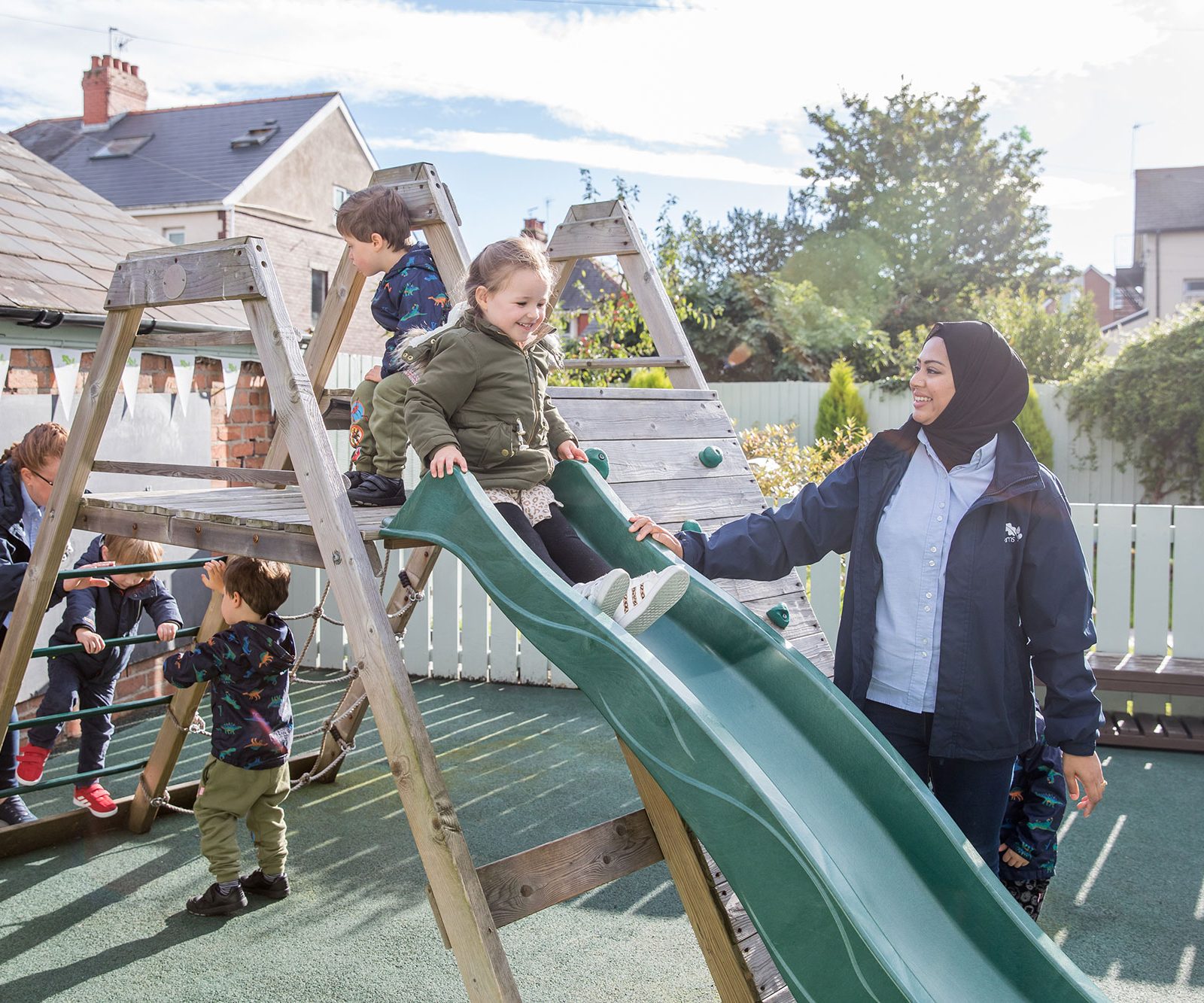 Acorns Nurseries Children and caregivers enjoy a sunny day on the playground slide at Oakfield Street, with a climbing frame in the background surrounded by acorns.