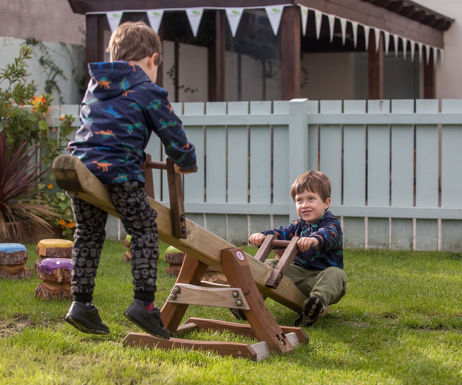 Acorns Nurseries Two children play on a wooden seesaw in a grassy backyard on Oakfield Street, with a white fence and bunting in the background. Acorns scatter around, adding a touch of autumn charm to their joyful afternoon.