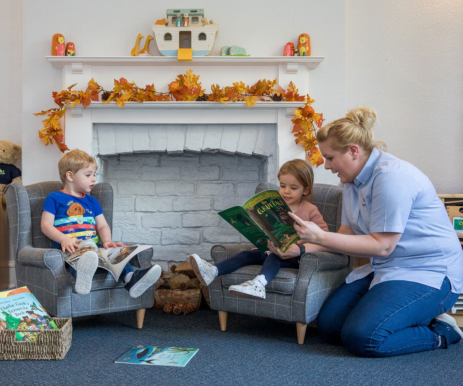 Acorns Nurseries A woman reads a book to a young boy and girl seated in a cozy playroom on Oakfield Street, where the fireplace is beautifully adorned with fall-themed decorations, featuring an array of colorful acorns.