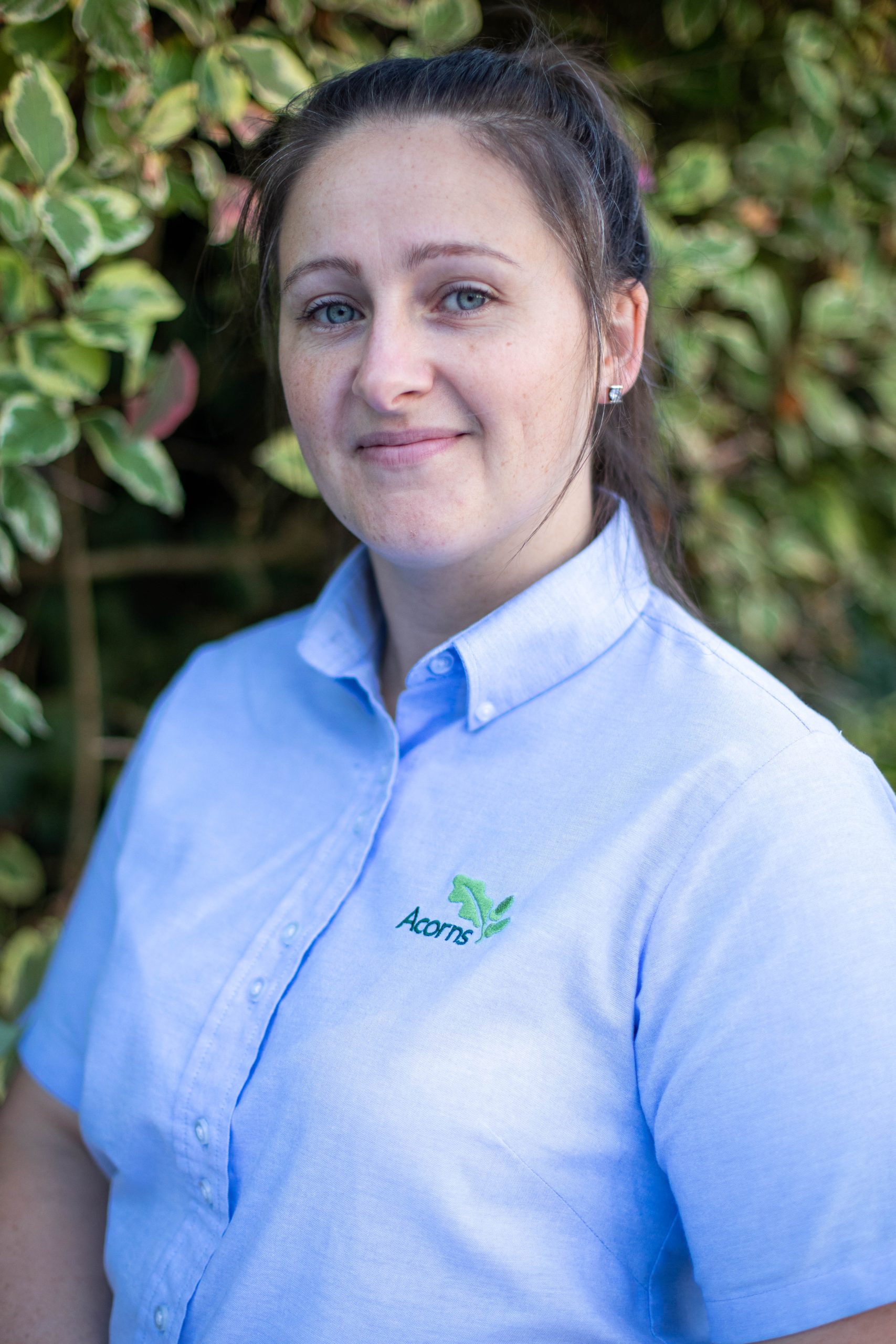 Acorns Nurseries A person in a light blue shirt with an Acorns ONS logo stands outside in front of green foliage, looking at the camera with a neutral expression, perhaps on a break from the Workplace Nursery.