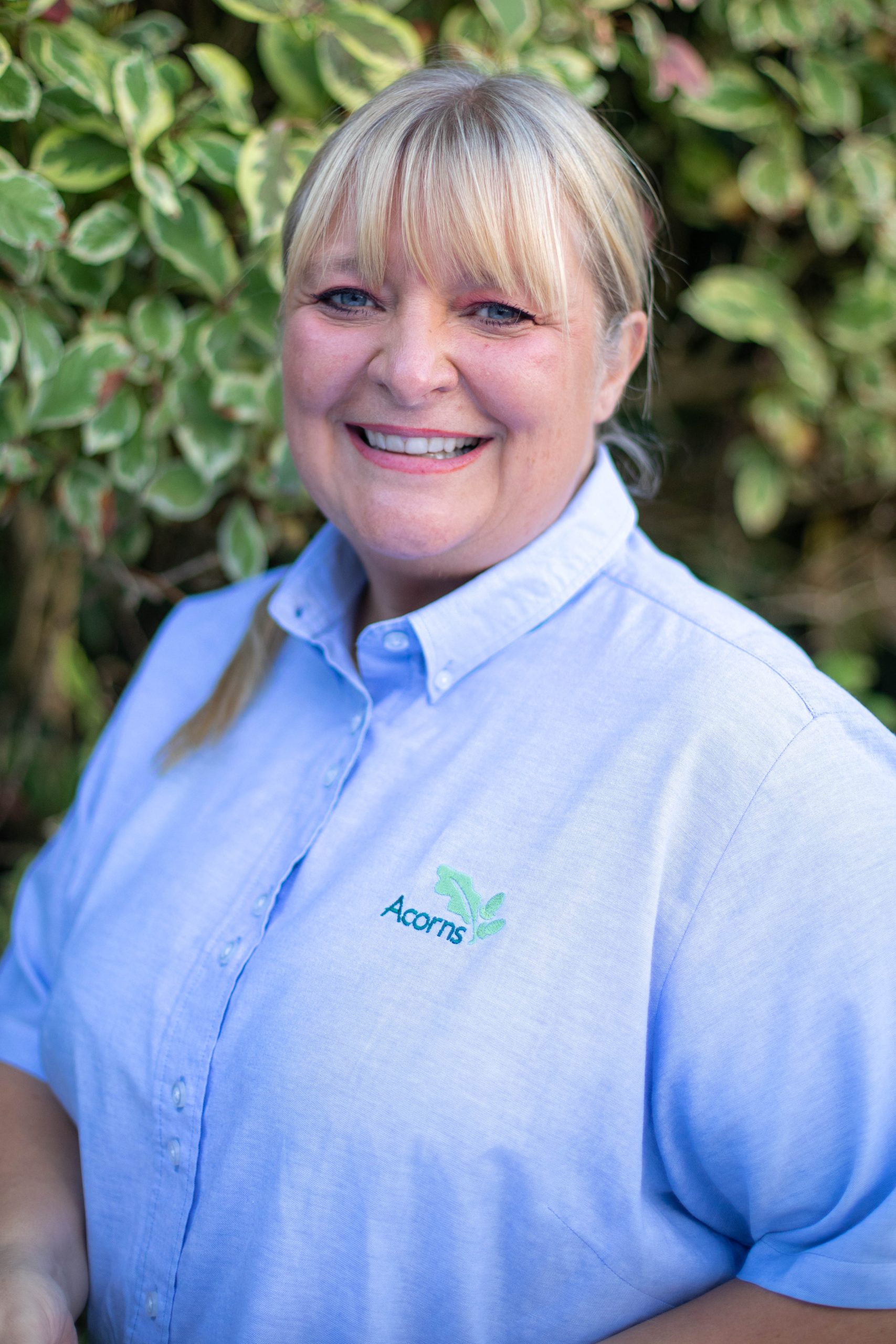 Acorns Nurseries A smiling person with blonde hair, wearing a light blue shirt adorned with an acorn logo, stands gracefully in front of the lush greenery of Parc Nantgarw.