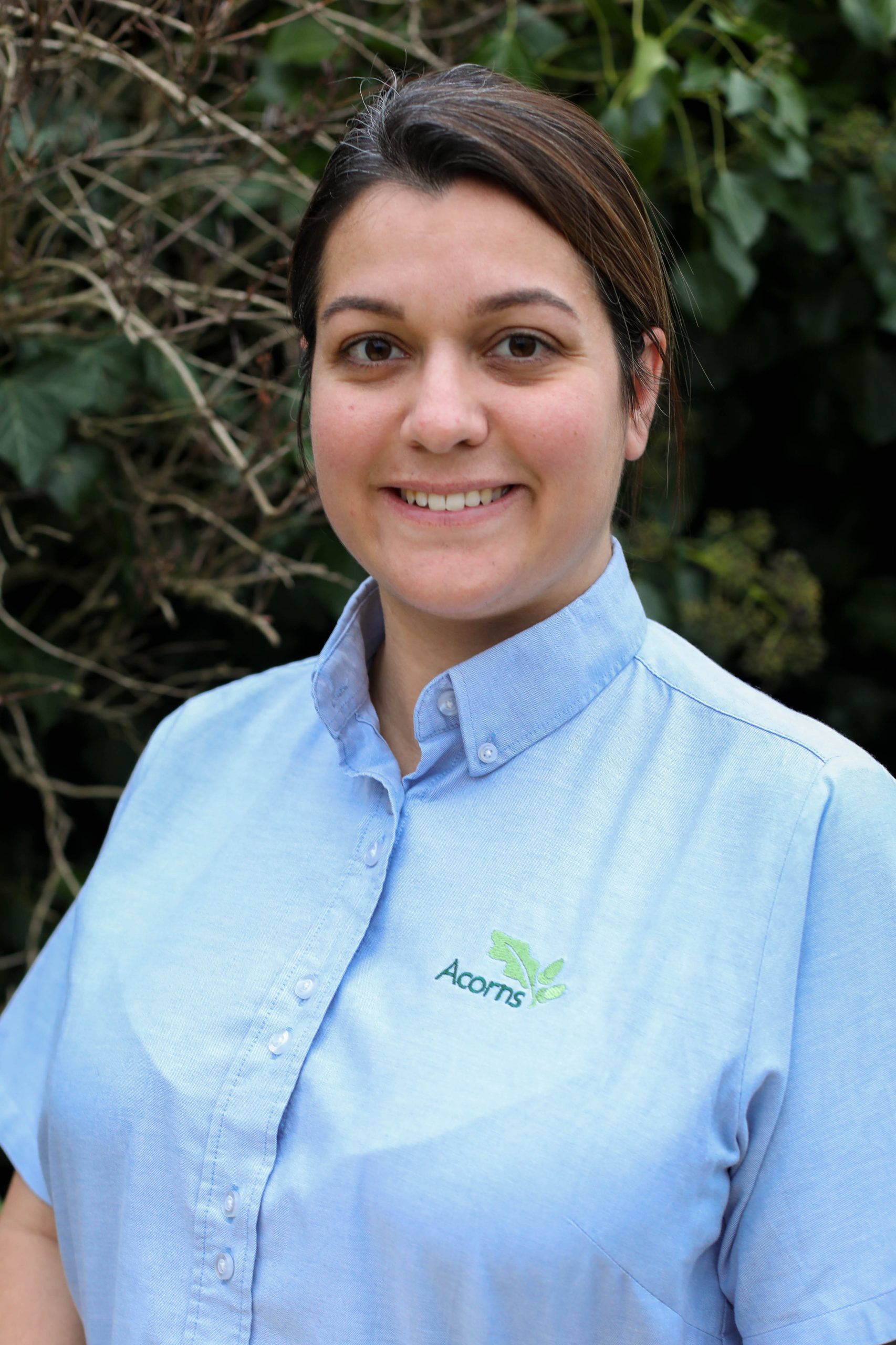 Acorns Nurseries A person wearing a blue Acorns shirt beams with joy outdoors, surrounded by lush greenery.