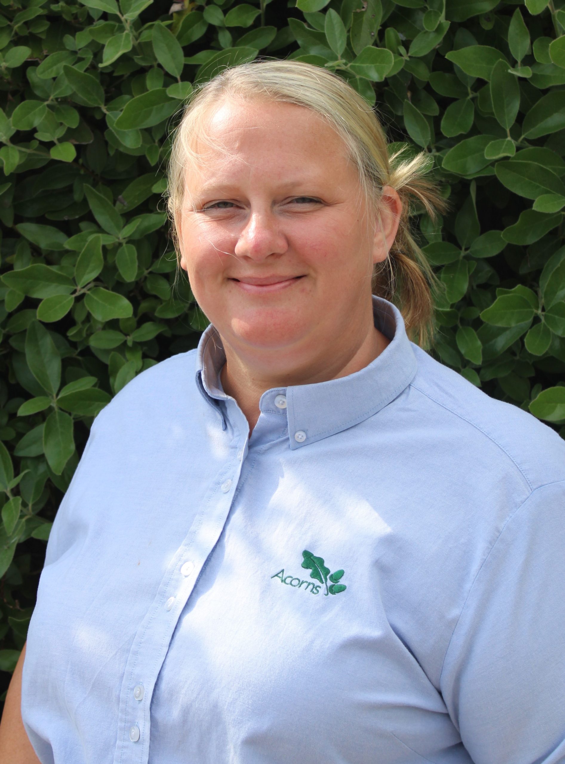 Acorns Nurseries A person in a blue shirt with an Acorns logo stands in front of a leafy green background at Parc Nantgarw, smiling at the camera.