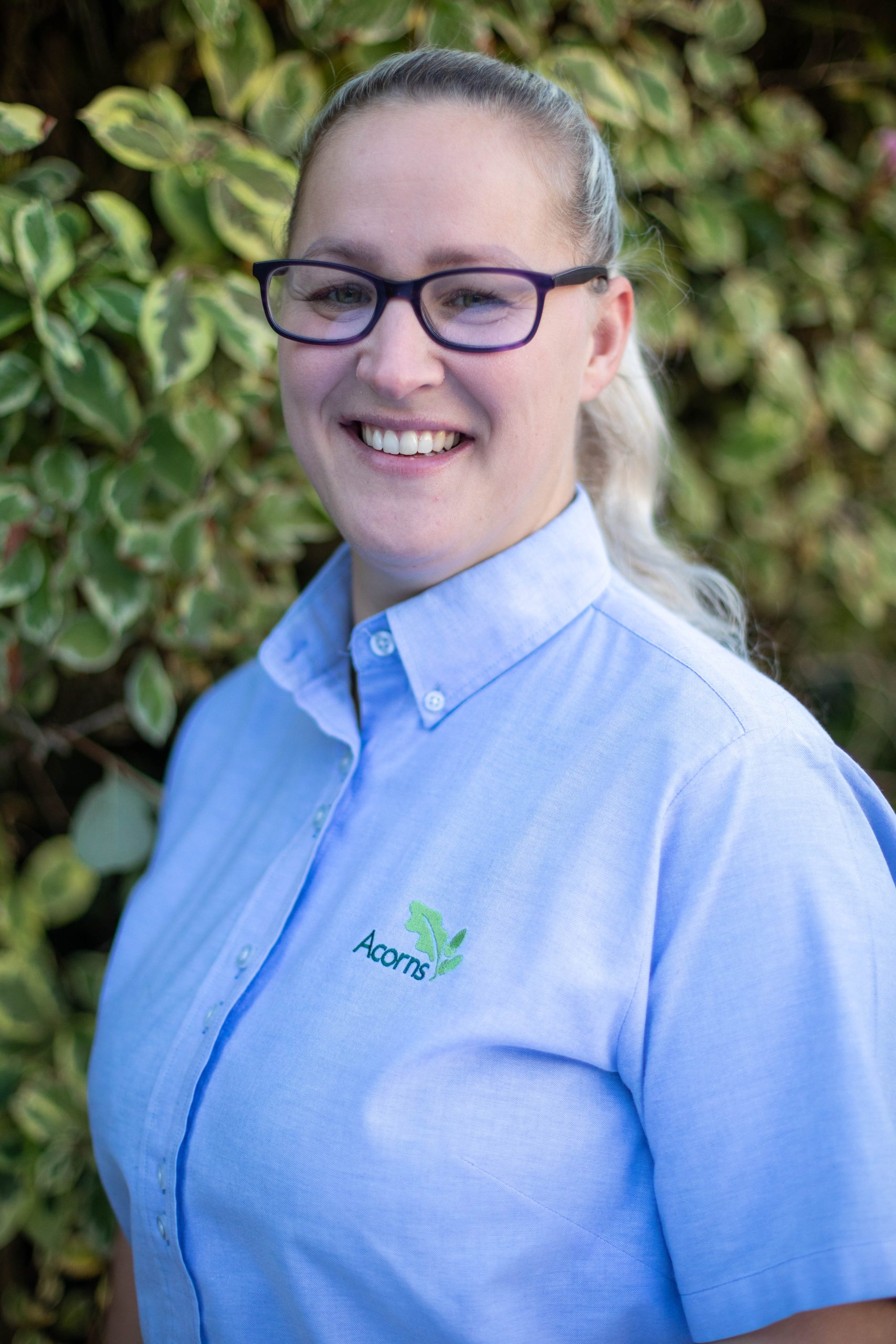 Acorns Nurseries A smiling person with glasses, wearing a blue Henbury Hill House shirt featuring the 