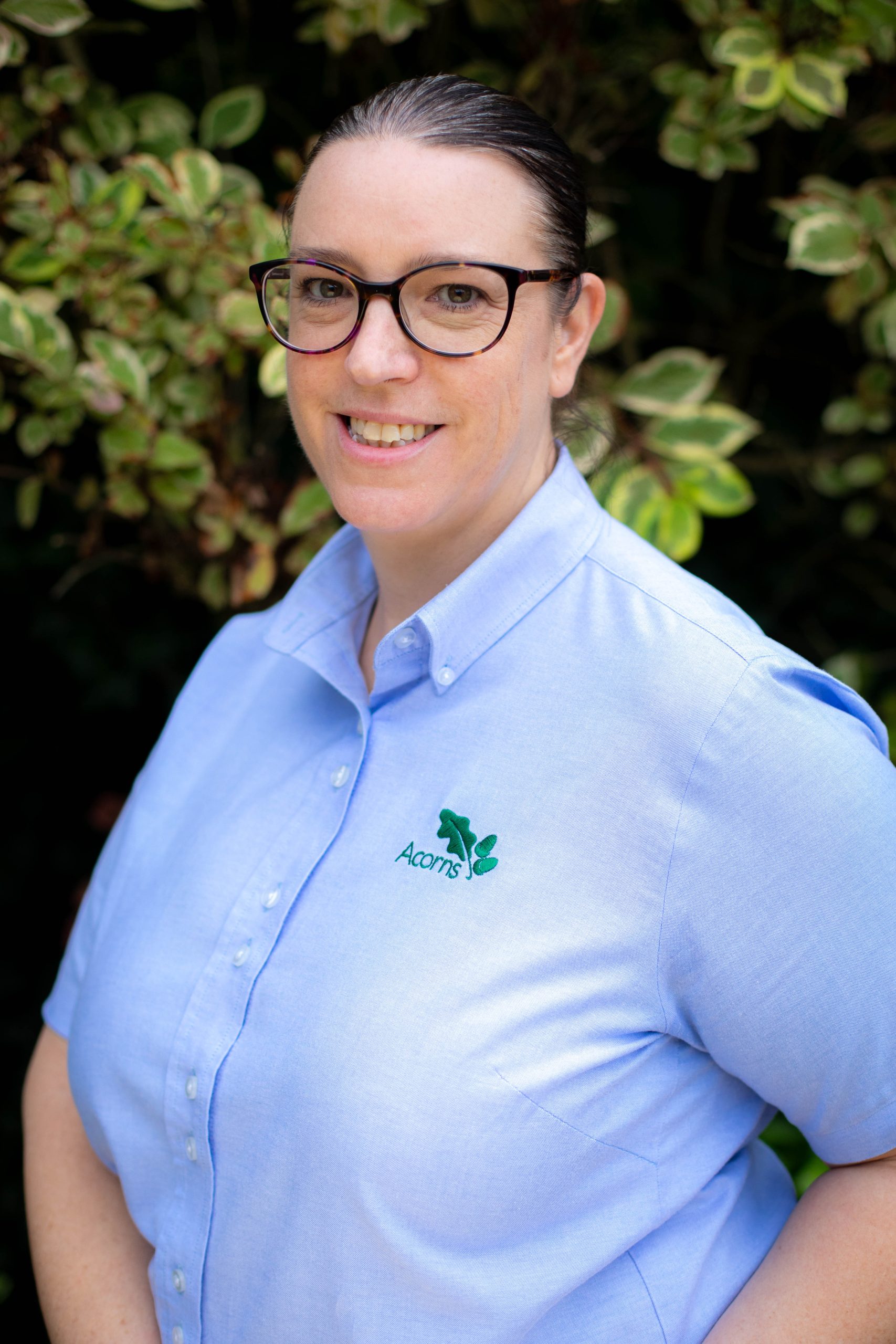 Acorns Nurseries Standing outdoors in front of leafy greenery, a person wearing glasses and a light blue shirt with an Acorns logo smiles warmly at the camera, reminiscent of a sunny day in Llandaff.