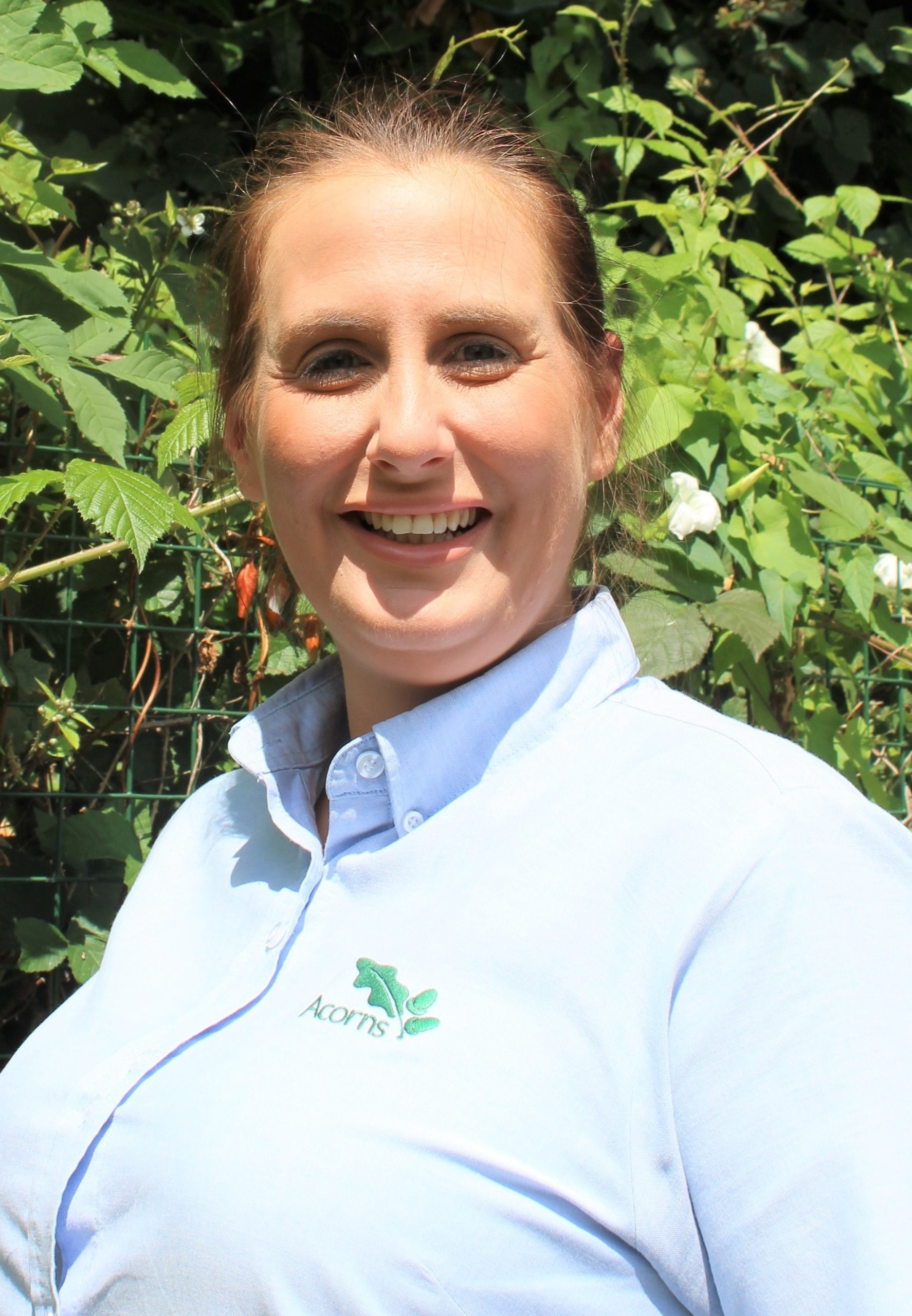 Acorns Nurseries A smiling person in a light blue shirt stands outdoors at Duffryn, surrounded by lush green foliage and scattered acorns in the background.