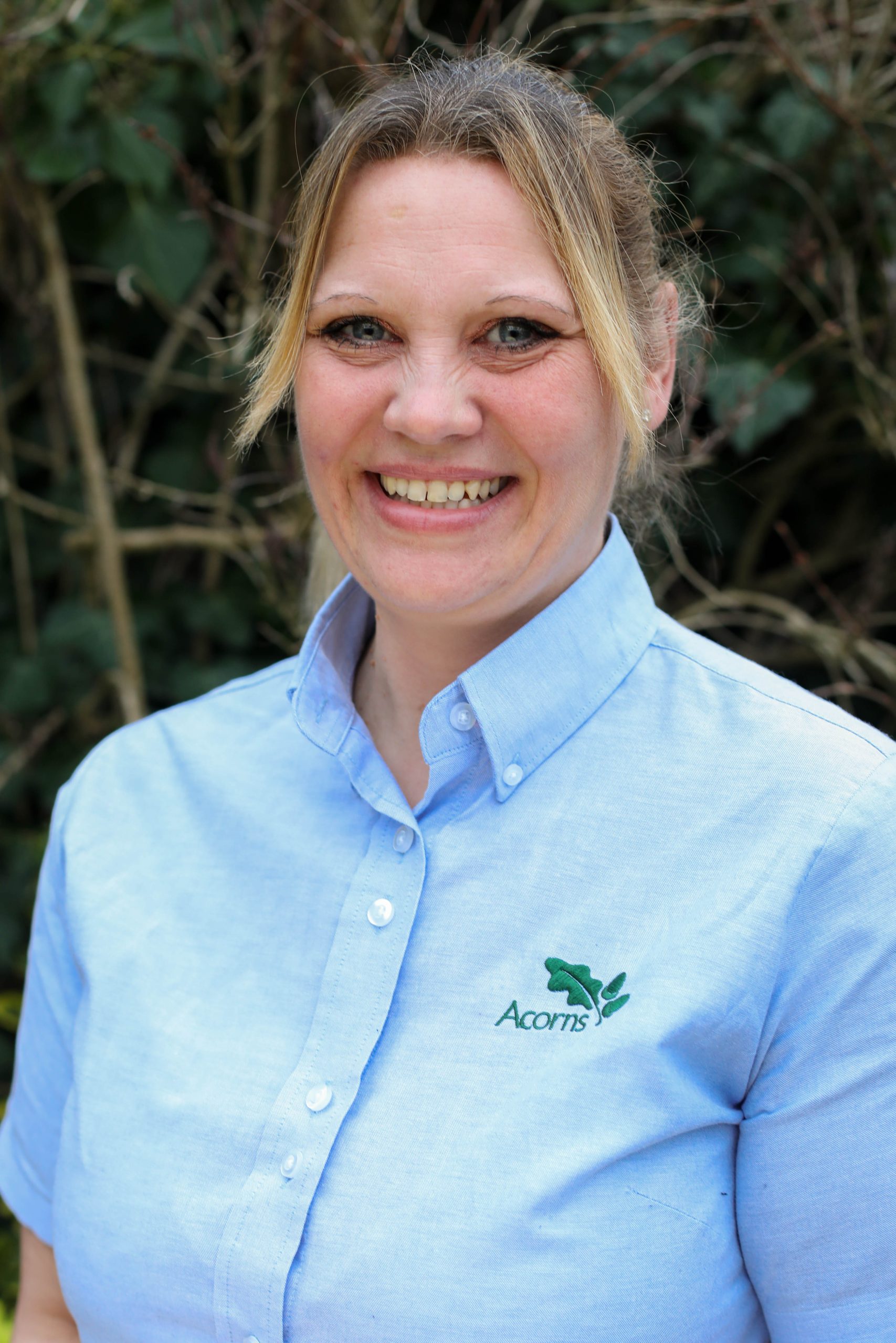 Acorns Nurseries A smiling person in a light blue shirt stands outdoors in front of the lush greenery of Henbury Hill House, where vibrant acorns dapple the ground.