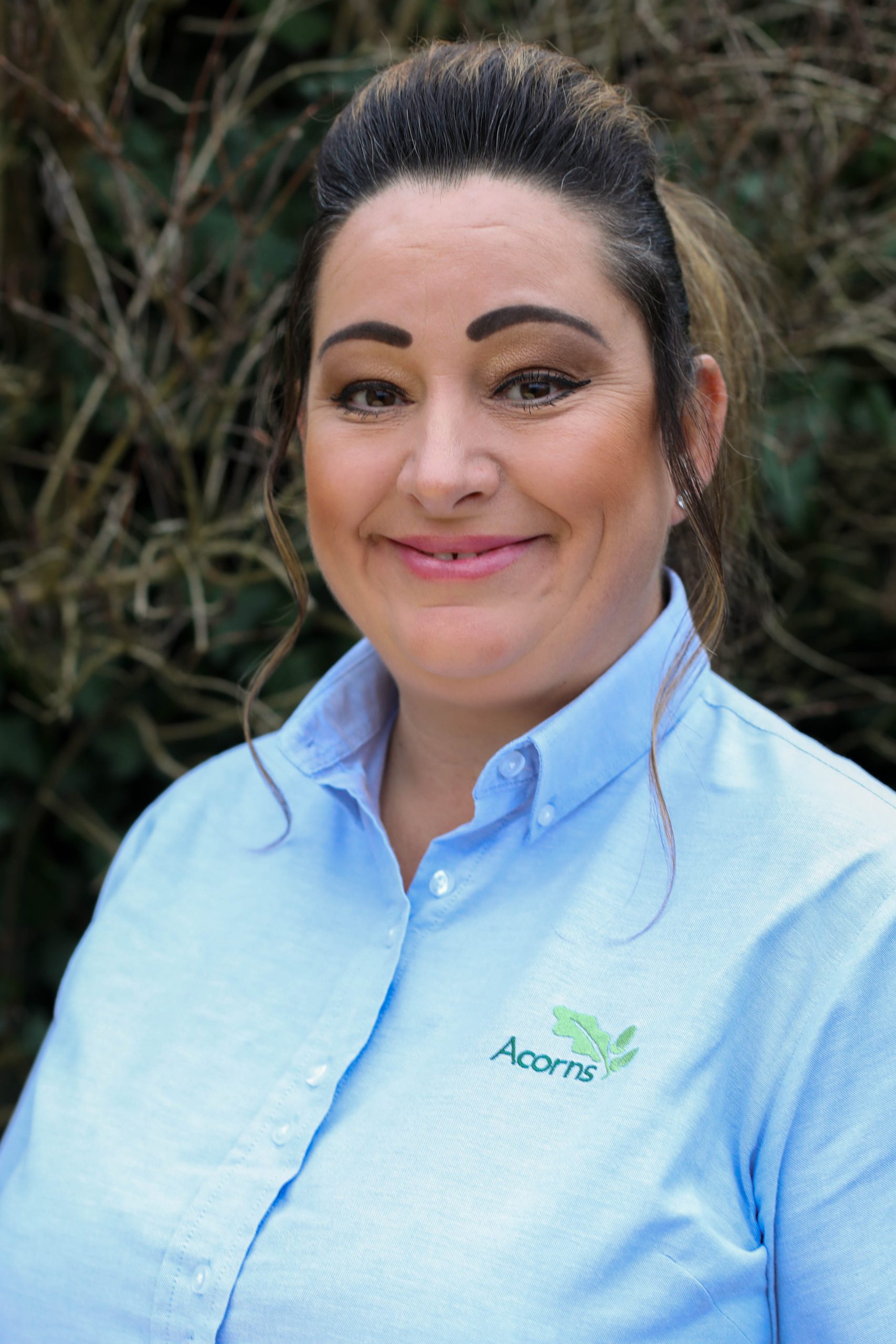 Acorns Nurseries A woman in a blue Acorns shirt stands outdoors, smiling warmly amidst a backdrop of twigs and greenery, embodying the spirit of growth and nature.