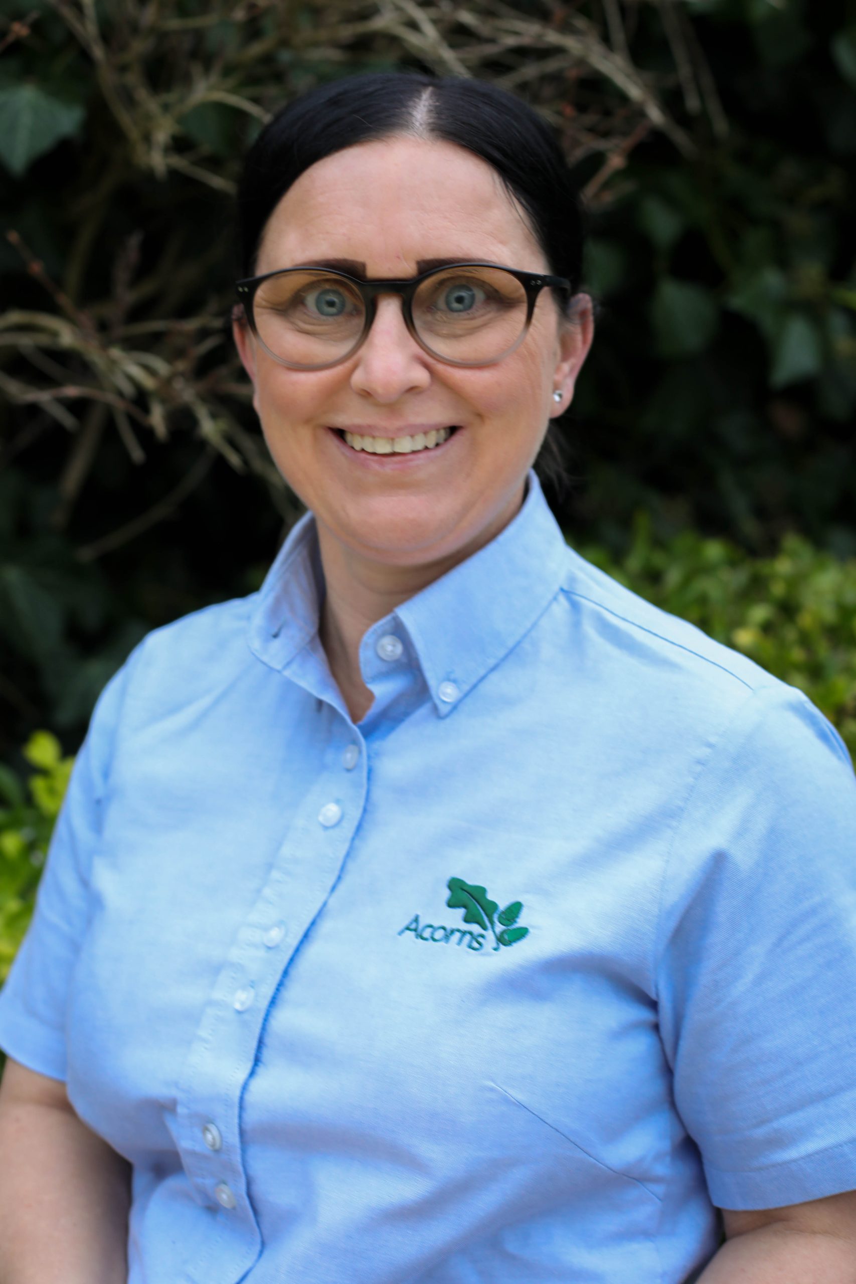 Acorns Nurseries A person with glasses smiles, donning a light blue shirt emblazoned with the Acorns logo, standing amidst lush greenery.