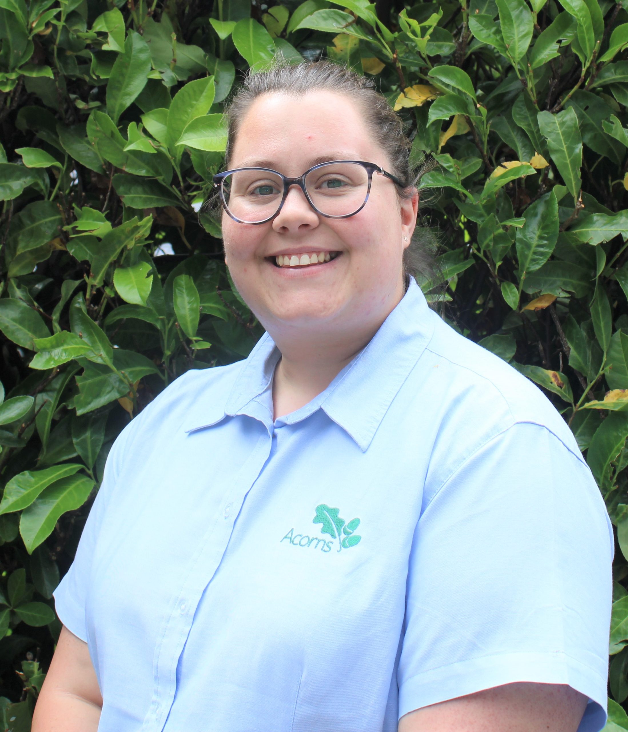 Acorns Nurseries A person wearing glasses and a blue shirt with an embroidered logo stands in front of the lush green foliage of Henbury Hill, smiling at the camera.