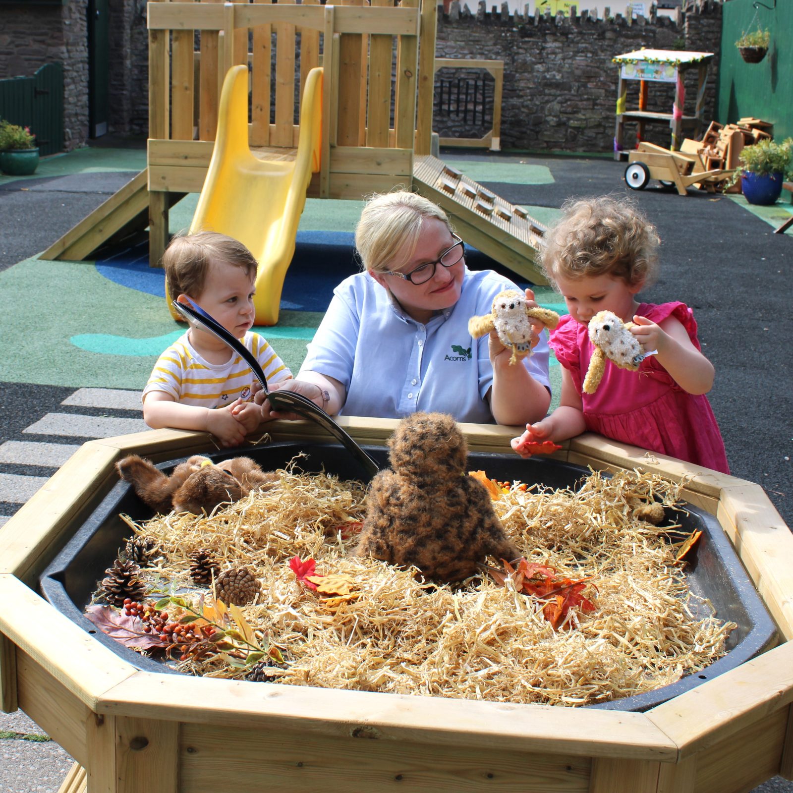 Acorns Nurseries At Acorns House, two children and an adult delight in the playful wonder of toy owls at a sensory play table in the playground.