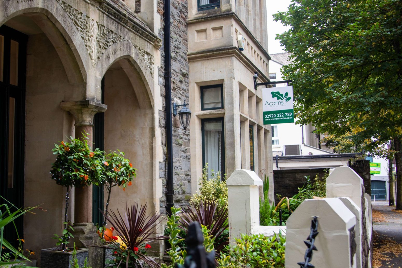 Acorns Nurseries A charming stone building with arched details stands prominently, its front garden enhancing the allure of Acorns House. Trees line the sidewalk, creating a picturesque setting for this inviting abode.