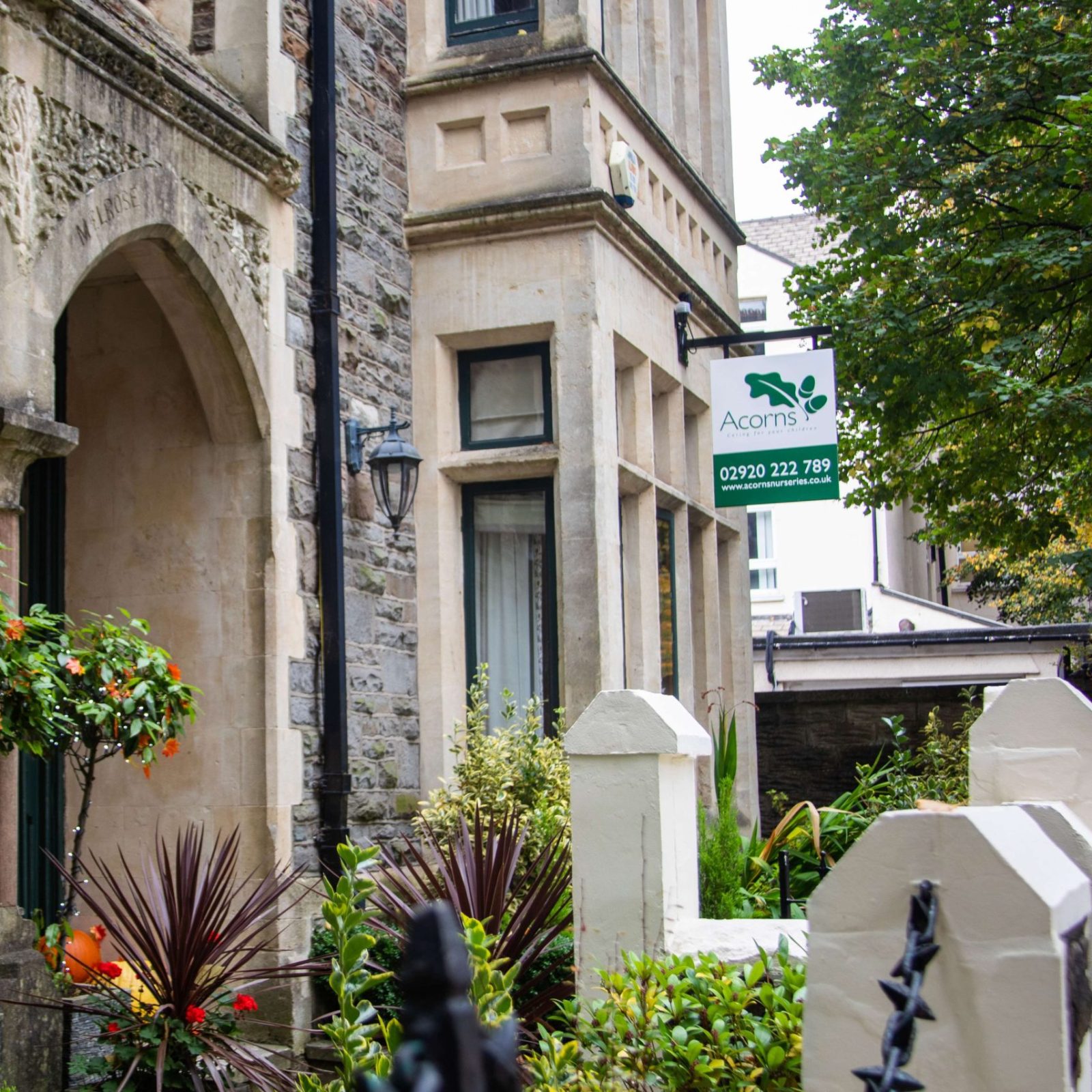 Acorns Nurseries A charming stone building with arched details stands prominently, its front garden enhancing the allure of Acorns House. Trees line the sidewalk, creating a picturesque setting for this inviting abode.