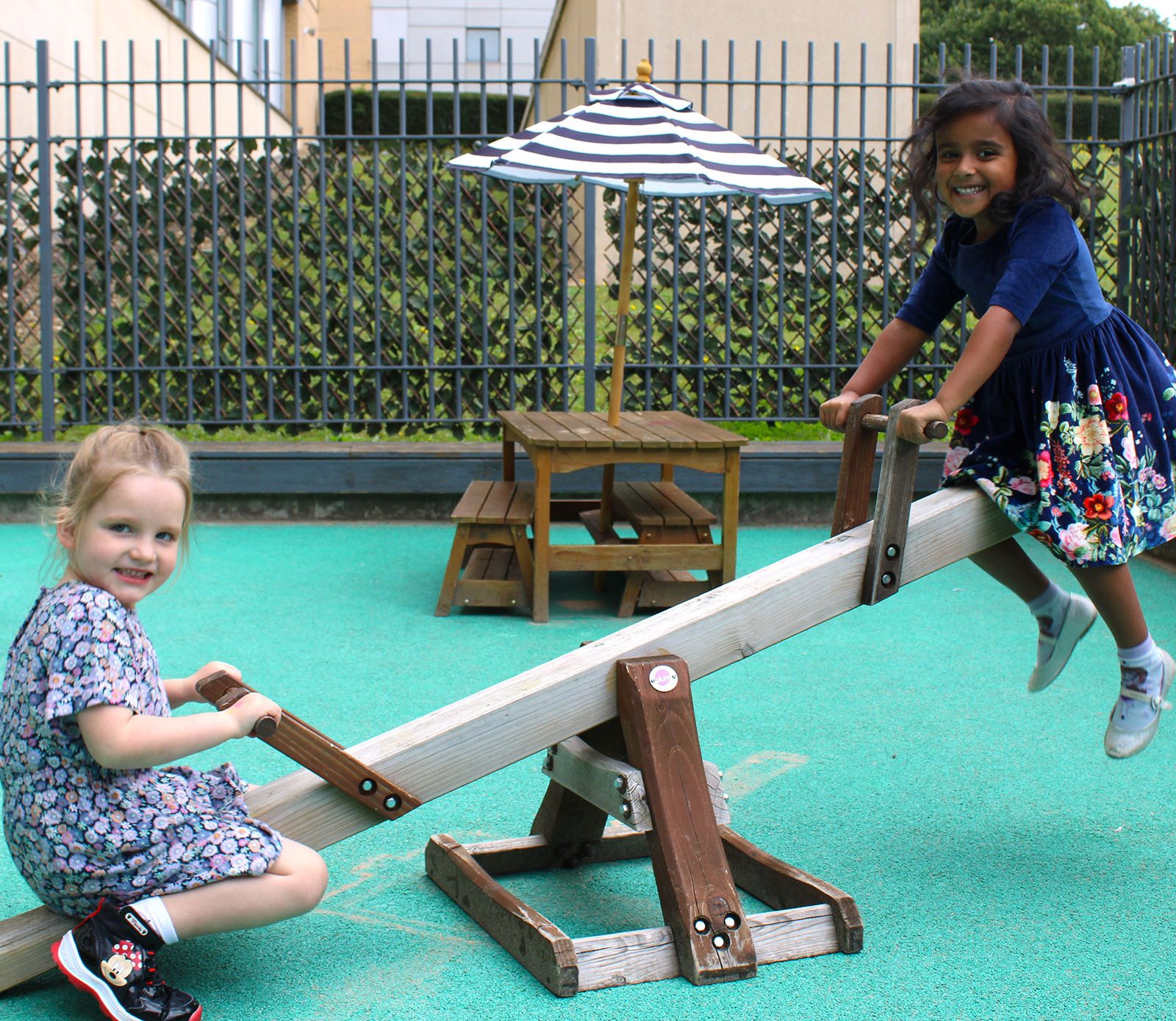 Acorns Nurseries Two children are joyfully playing on a seesaw at the Aviva-sponsored playground, where a picnic table and umbrella provide a picturesque backdrop.