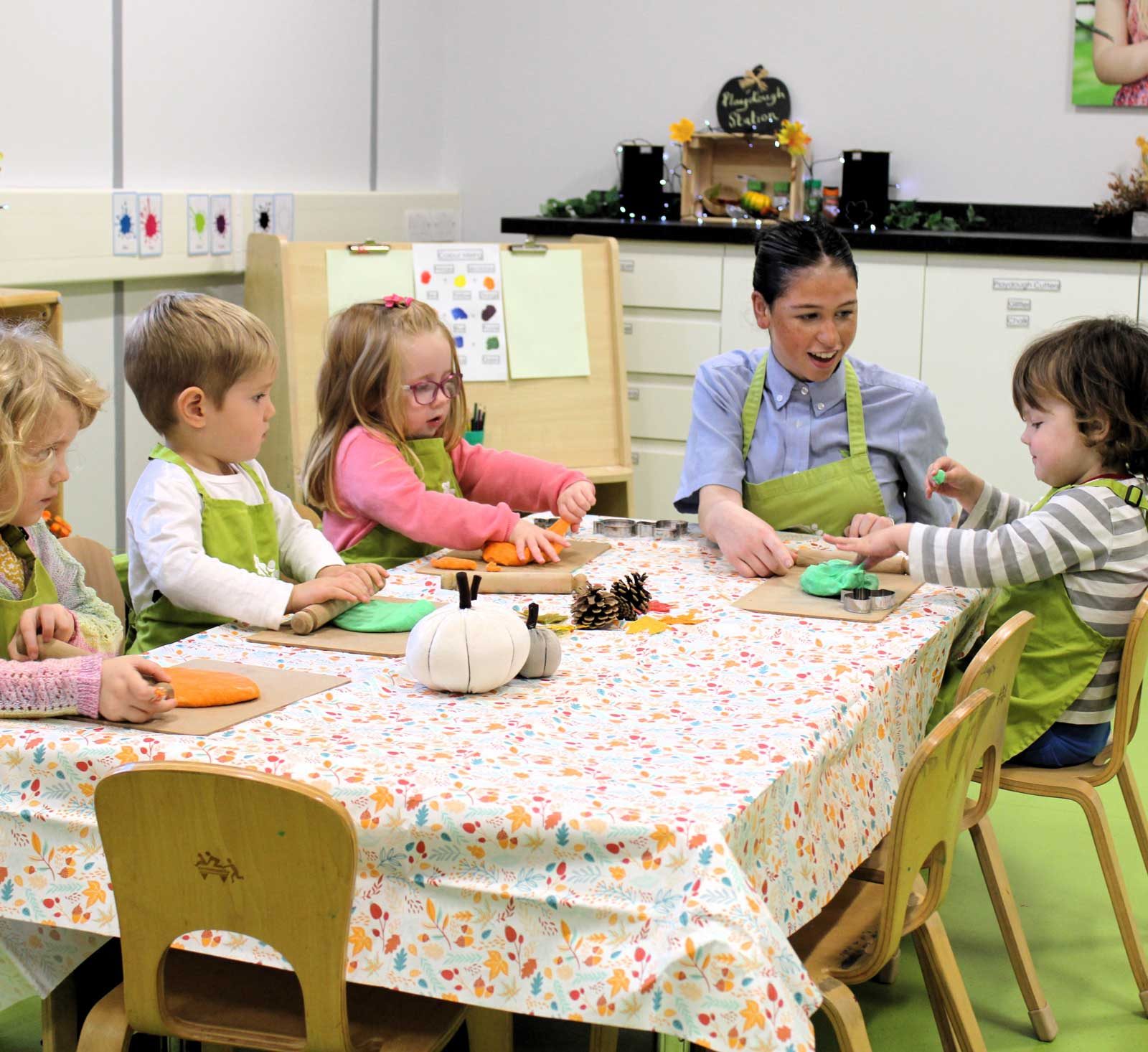 Acorns Nurseries In a lively workplace nursery, children and a teacher sit at a table, shaping colorful play dough into tiny acorns, delighting in their creative craft session.