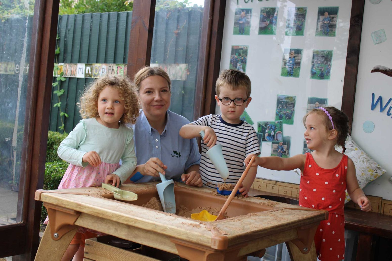 Acorns Nurseries At Oakfield Street, three children and a teacher are joyfully playing with sand around an indoor table, their giggles echoing like acorns rustling in the breeze.