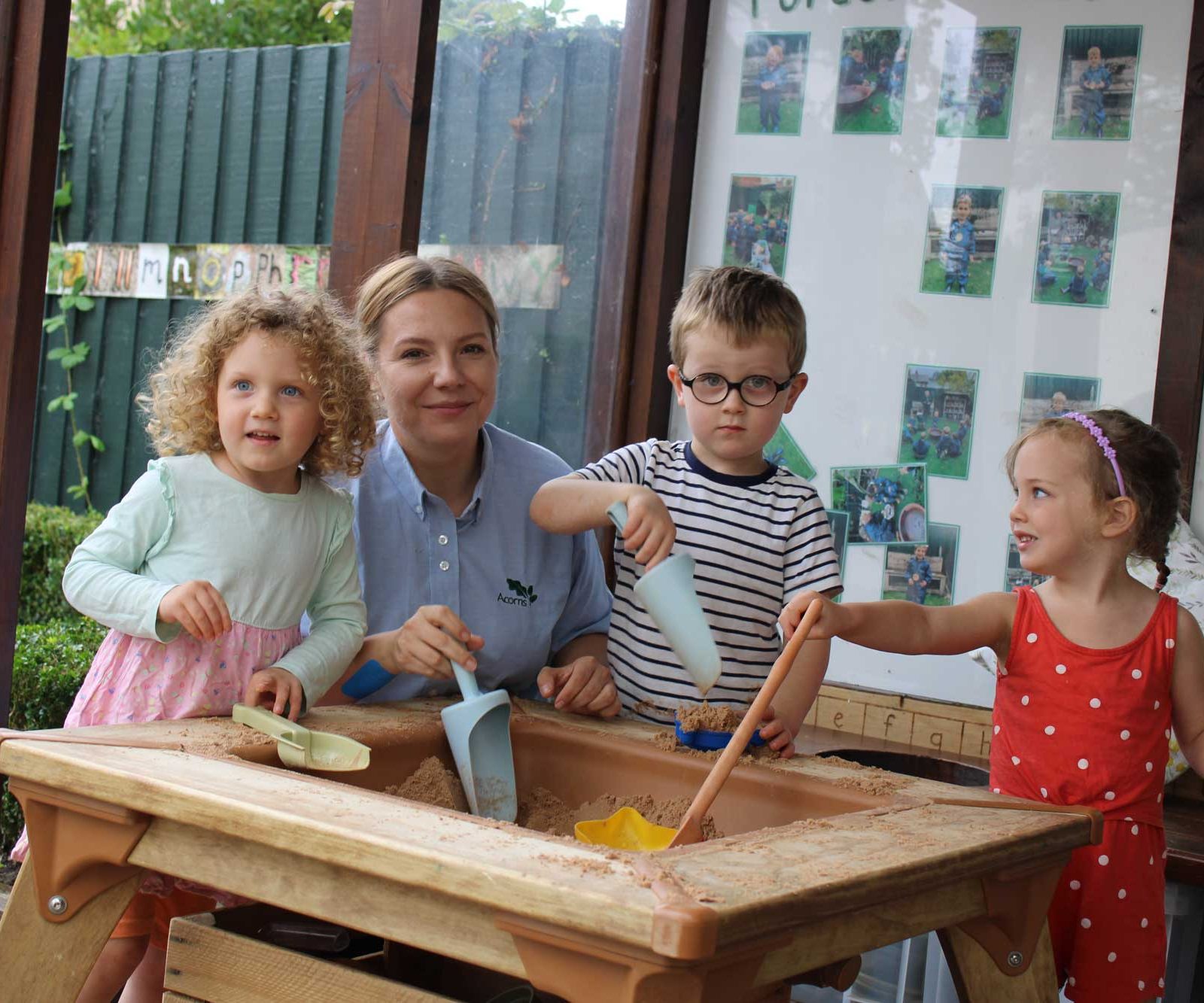 Acorns Nurseries At Oakfield Street, three children and a teacher are joyfully playing with sand around an indoor table, their giggles echoing like acorns rustling in the breeze.