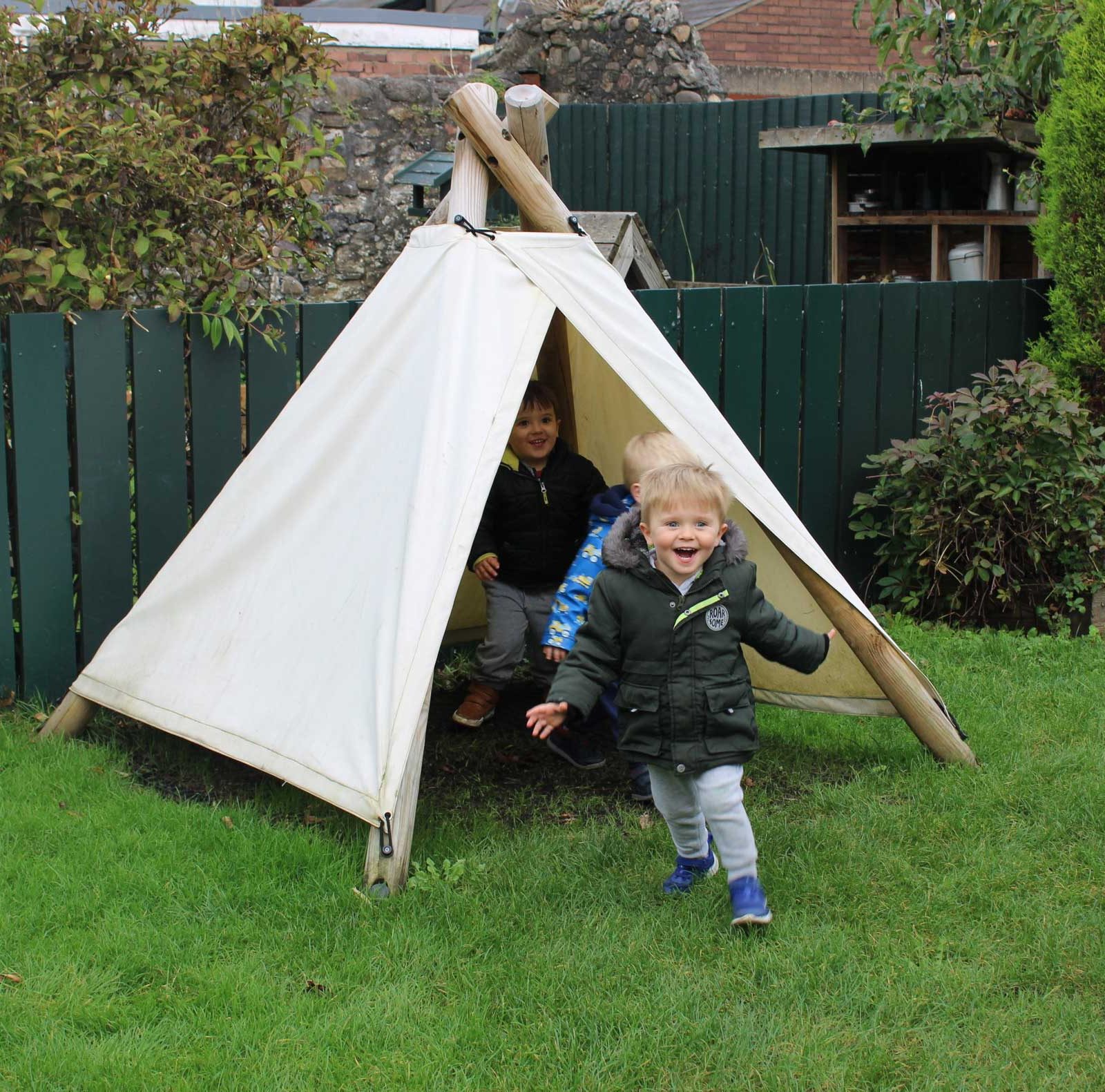 Acorns Nurseries Two children play in and around a small teepee in a grassy backyard on Oakfield Street. A wooden fence and shrubbery are in the background, with acorns scattered playfully across the yard.