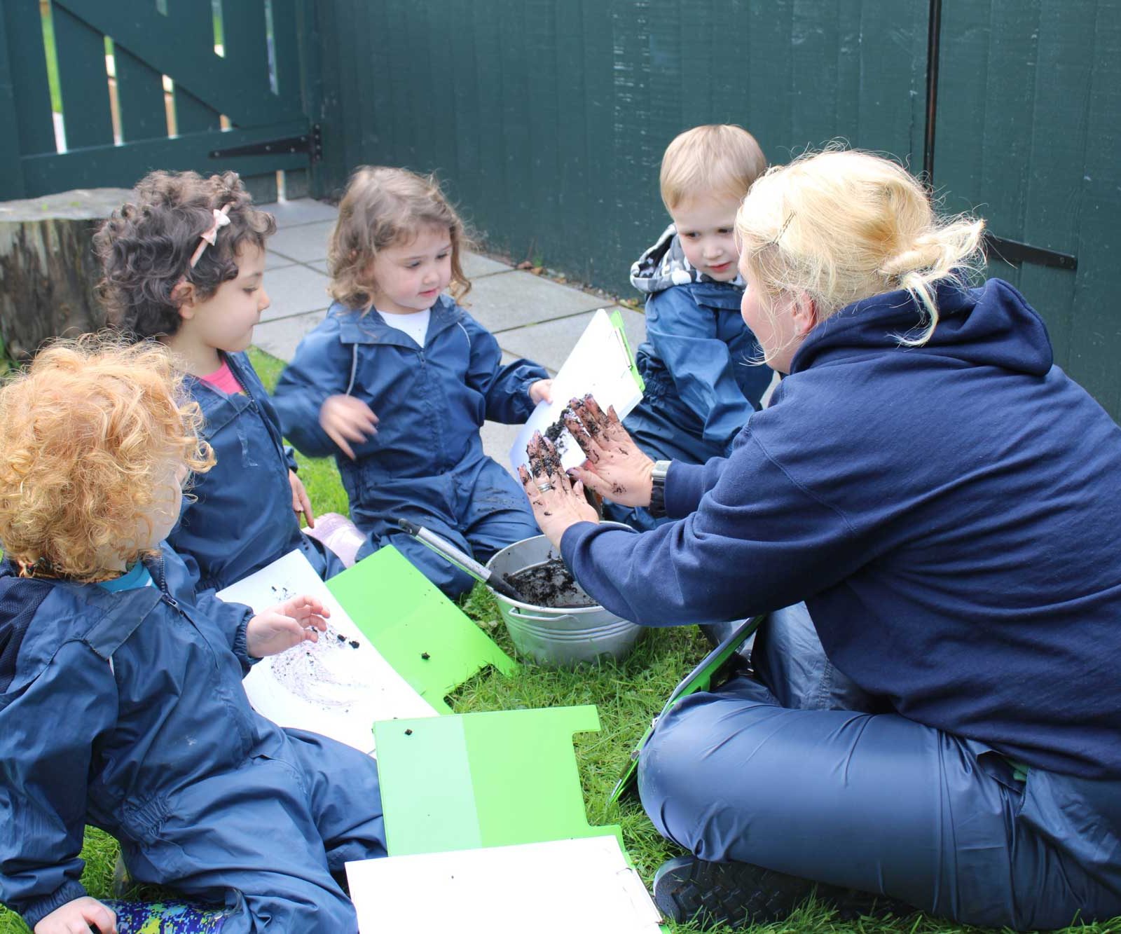 Acorns Nurseries A group of young children in rain suits gather around an adult examining soil samples under the towering Oakfield trees on a grassy area.