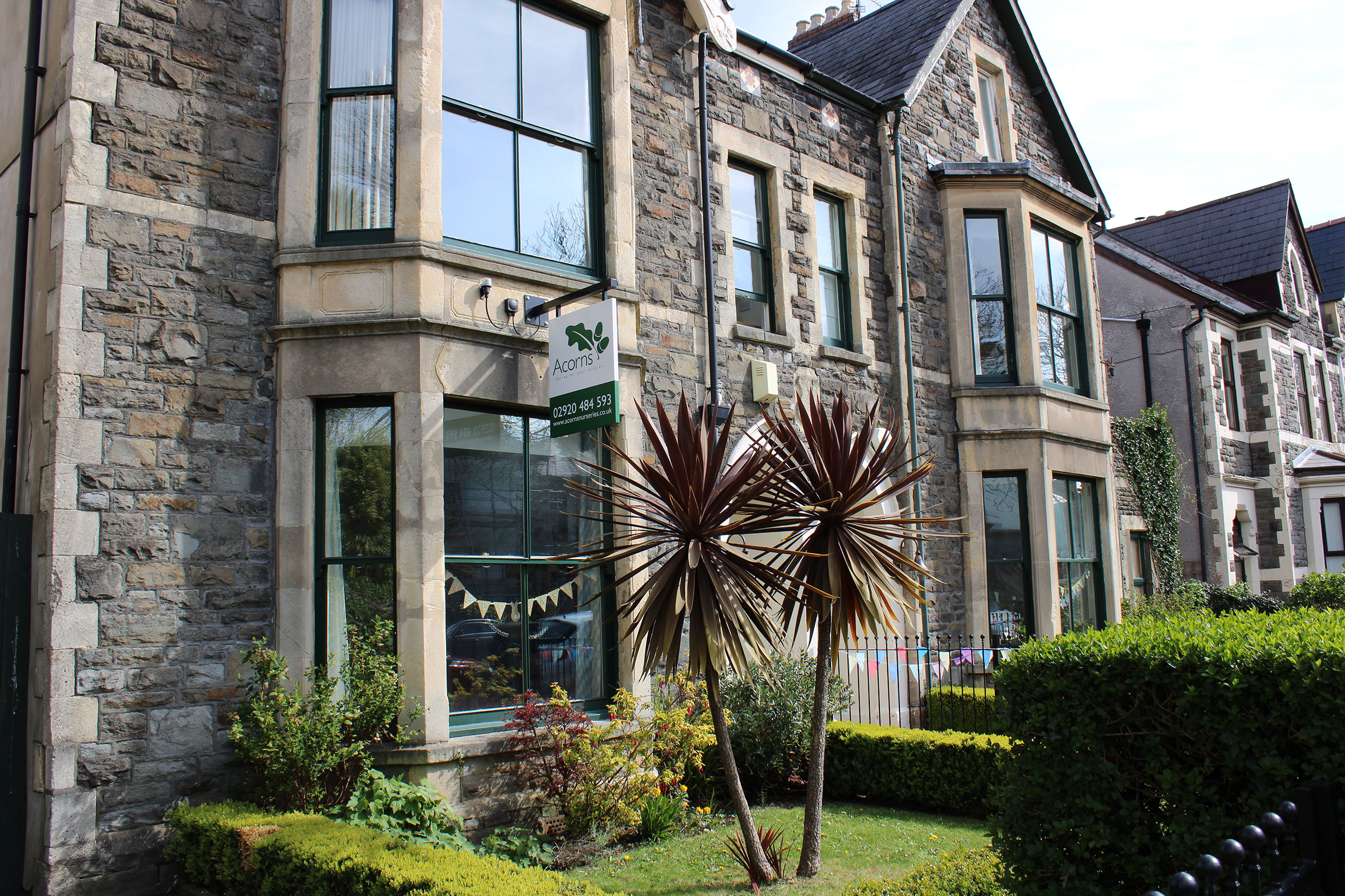 Acorns Nurseries A row of stone-built houses on Oakfield Street features charming bay windows and a small palm tree in the front yard, complete with a "For Sale" sign. A scattering of acorns adds a quaint touch to the picturesque setting.