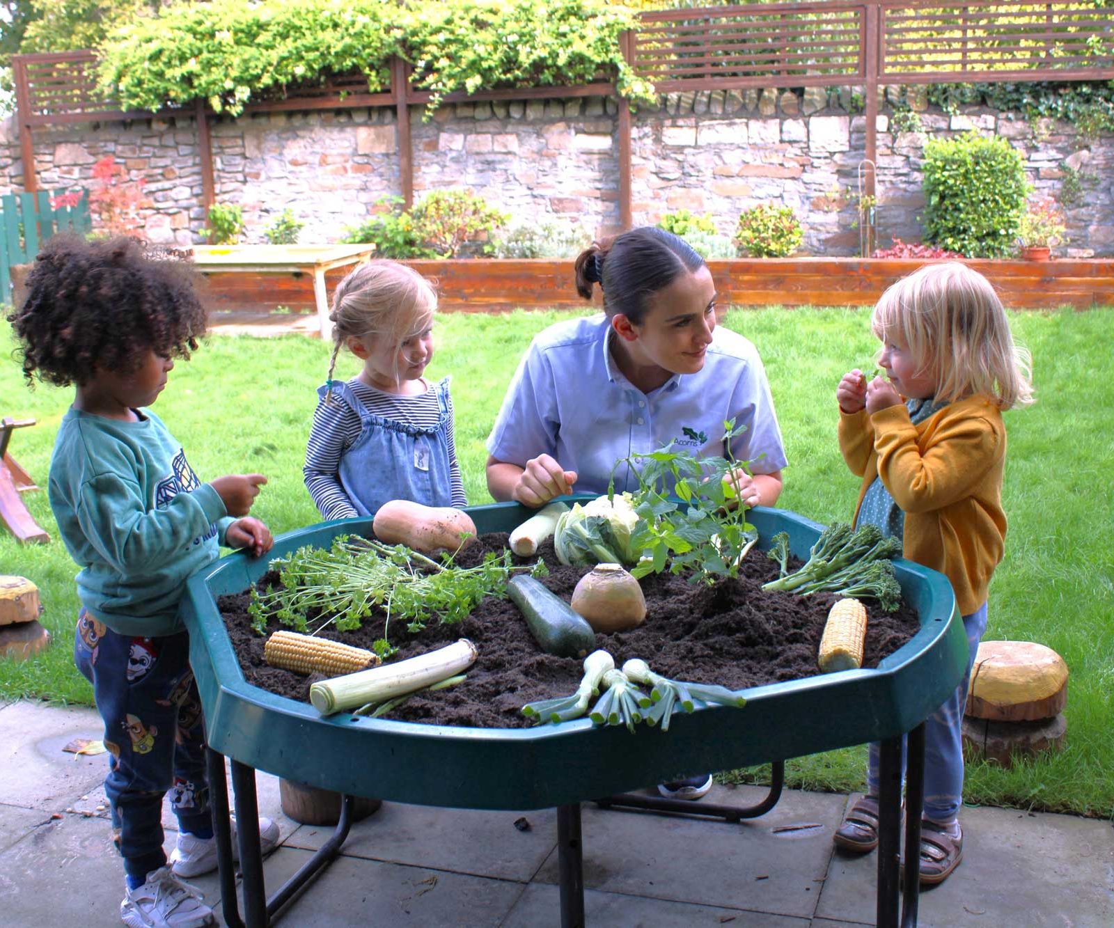 Acorns Nurseries In Whitchurch, children and an adult explore gardening at a table filled with soil, acorns, and various vegetables in an inviting outdoor setting.