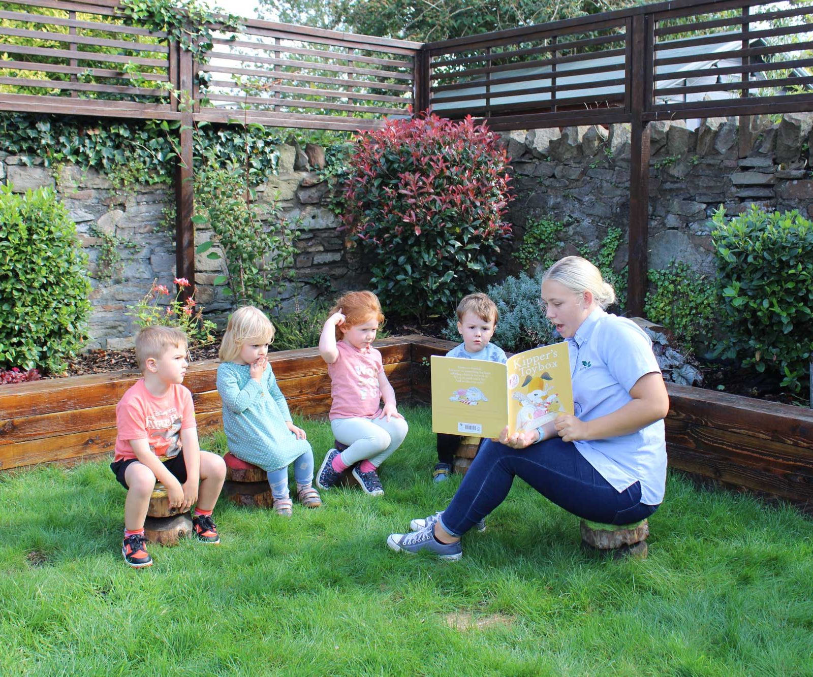 Acorns Nurseries In the serene garden setting of Whitchurch, a woman reads an enchanting book to four children seated on wooden stools, surrounded by lush greenery and a charming stone wall, like acorns nestled under a sturdy oak.