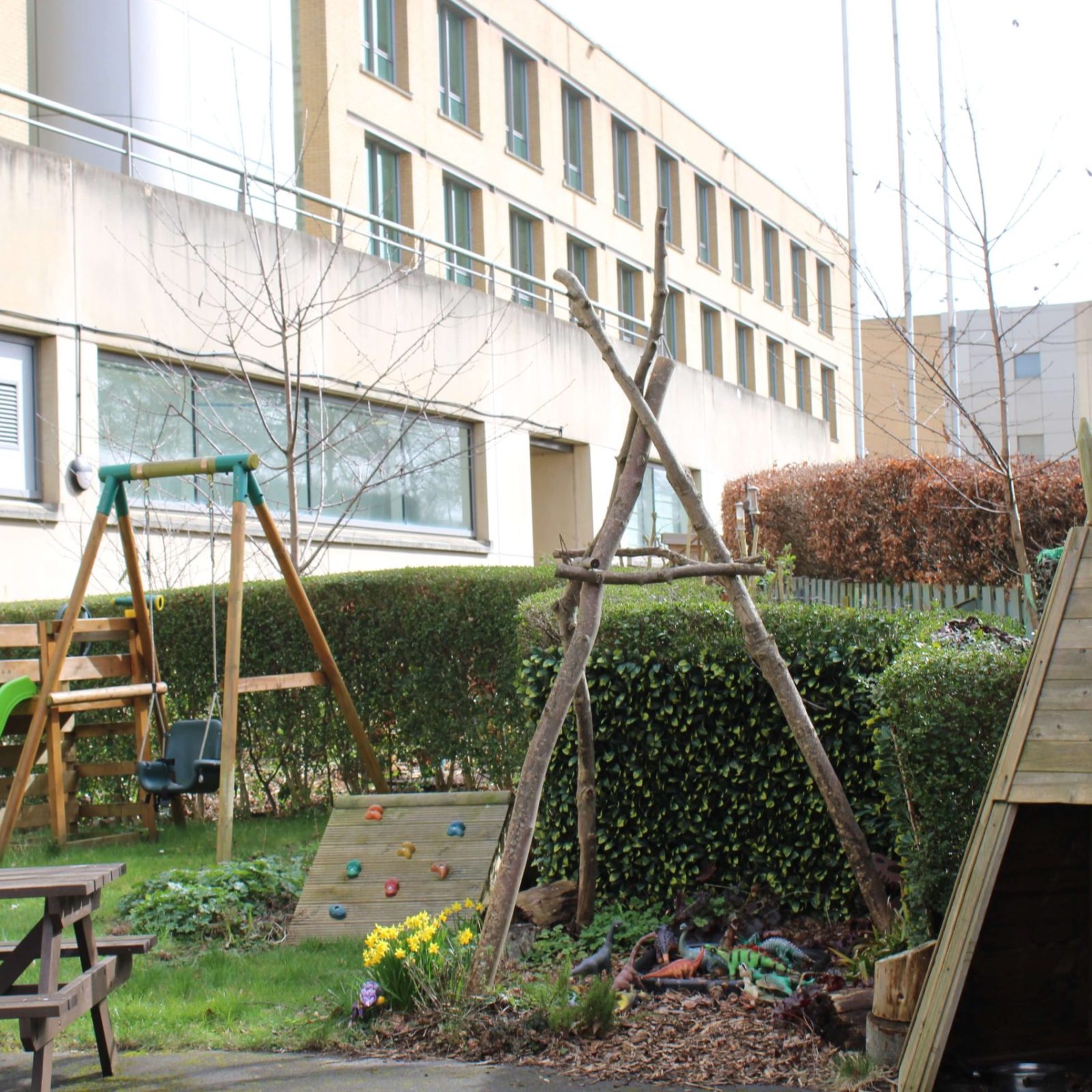 Acorns Nurseries The Acorns Aviva workplace nursery offers a delightful playground featuring a swing set, slide, climbing wall, and wooden structures nestled near a large building and lush hedges.