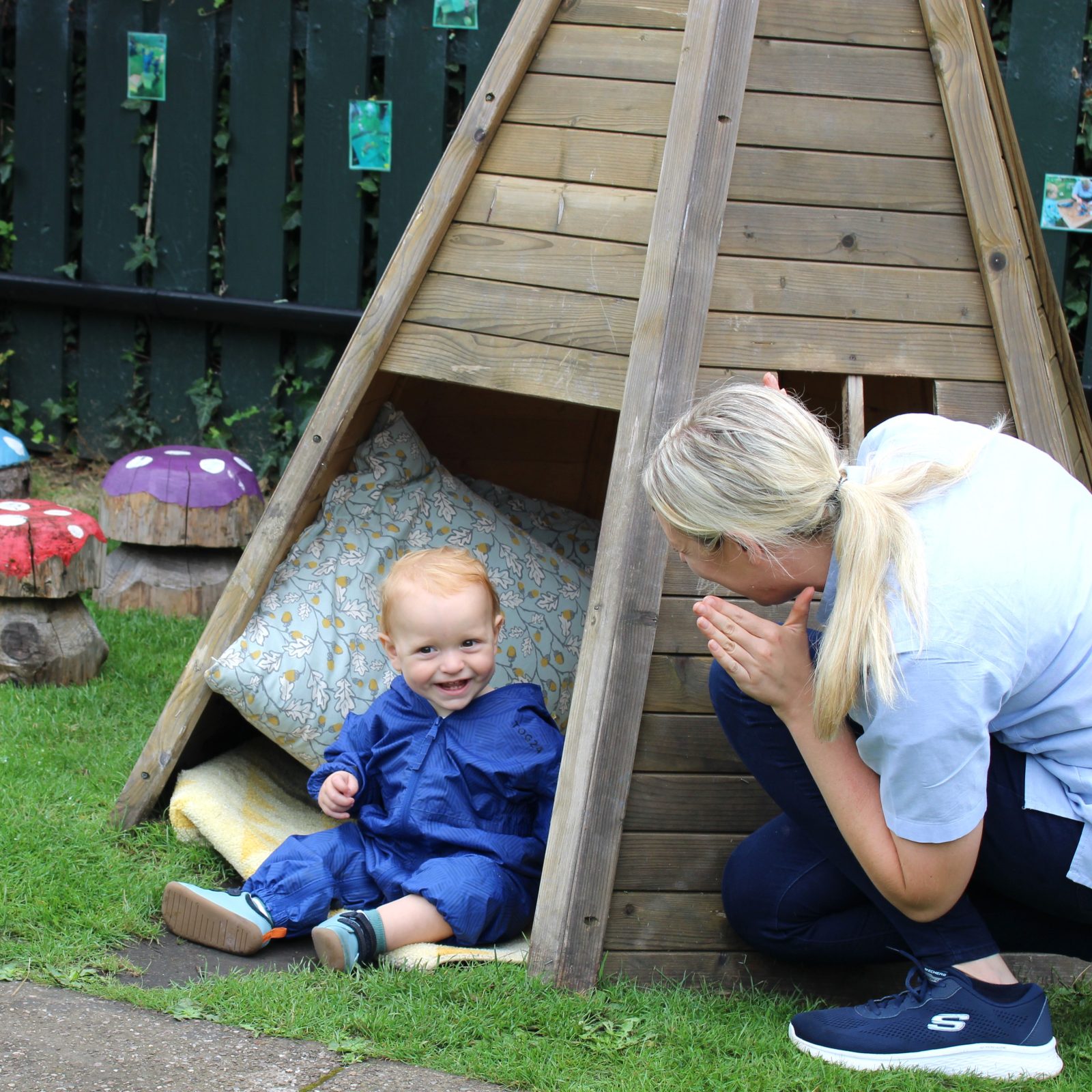 Acorns Nurseries A baby in a blue outfit sits in a wooden play structure, while an adult kneels nearby, facing the child with a smile.