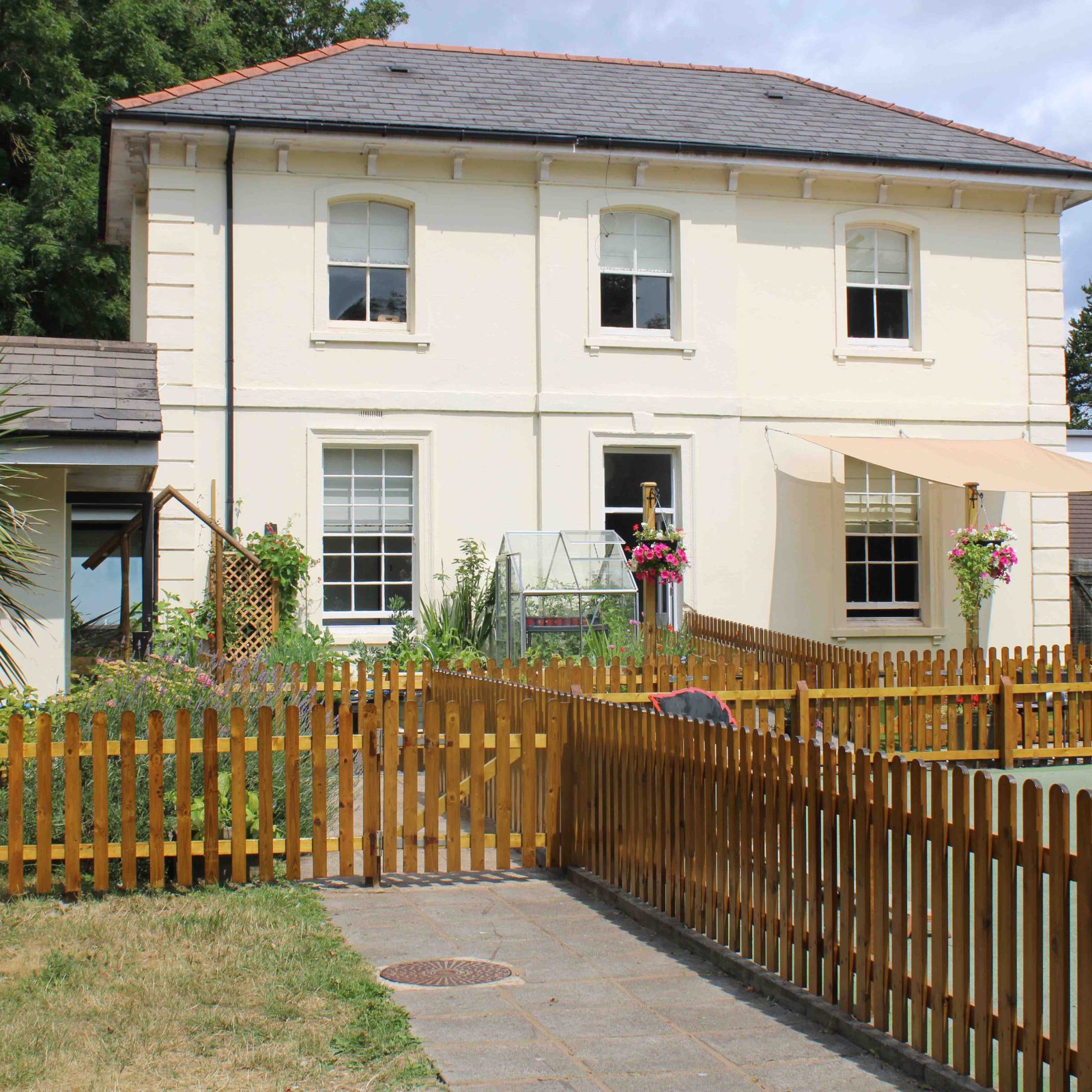 Acorns Nurseries A charming two-story cream house in Acorns Duffryn, with a gabled roof and surrounded by a wooden fence and garden. It features delightful awnings and outdoor decor, exuding a cozy ambiance.