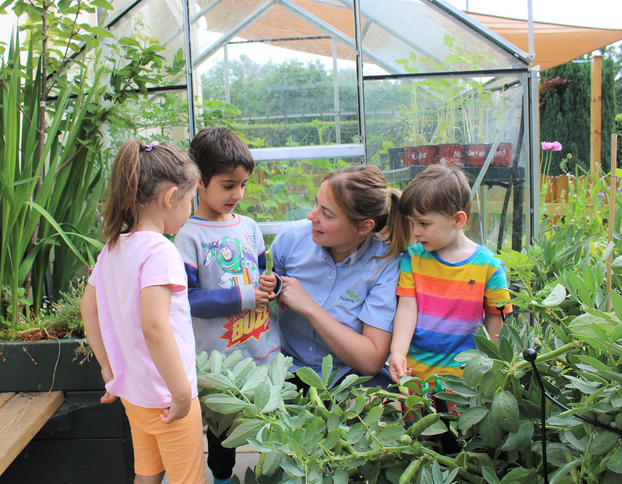 Acorns Nurseries In the lush garden near the greenhouse, a woman in a blue shirt engages three children in conversation, surrounded by nature's wonders—emphasizing the charm of Acorns Duffryn.