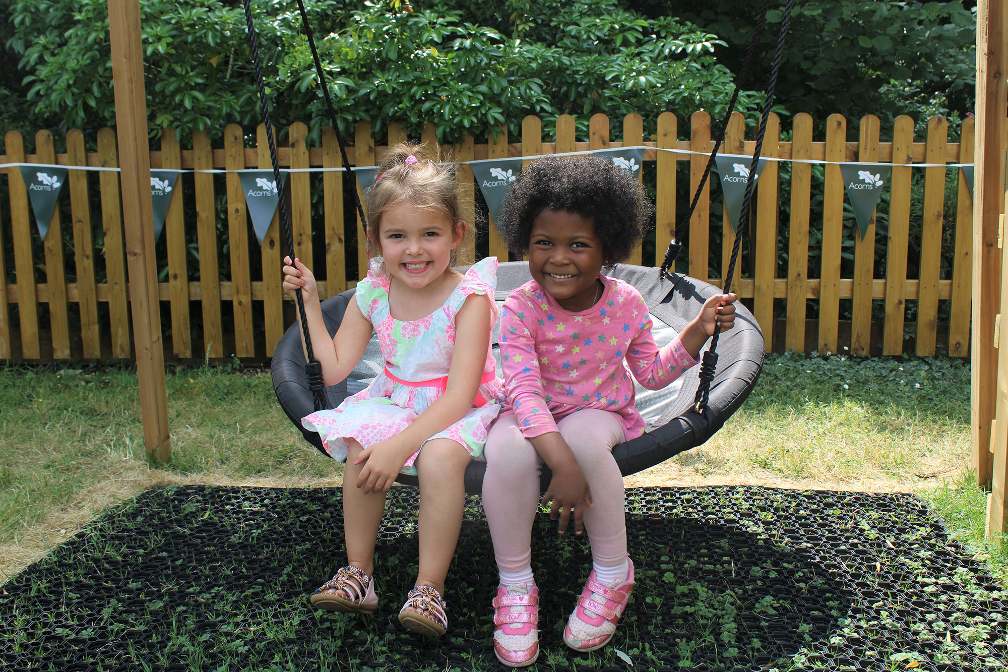 Acorns Nurseries Two young girls from Acorns Duffryn sit on a round swing, smiling brightly in a playground adorned with a wooden fence and lush greenery.