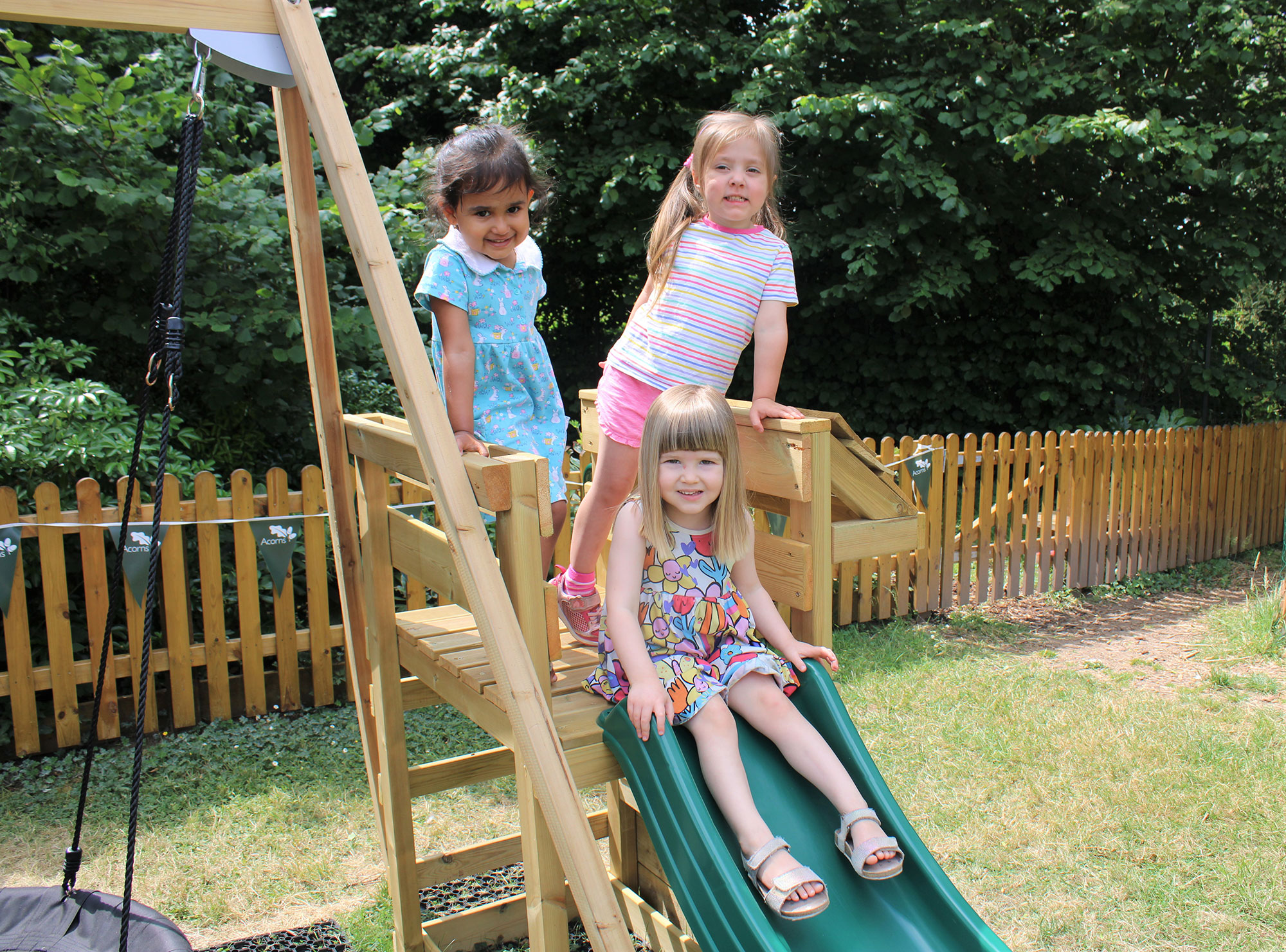 Acorns Nurseries Three children enjoy the Acorns Duffryn playground—a delightful wooden structure. Two of them stand proudly on the platform while one sits gleefully on the slide, surrounded by laughter and adventure.