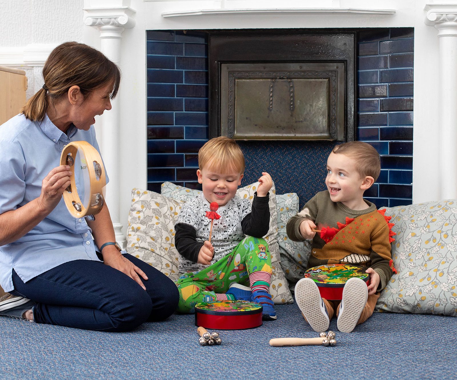 Acorns Nurseries In a cozy Whitchurch room with a blue-tiled fireplace and decorative pillows, an adult and two children sit on the floor, surrounded by acorns, joyfully playing musical instruments.