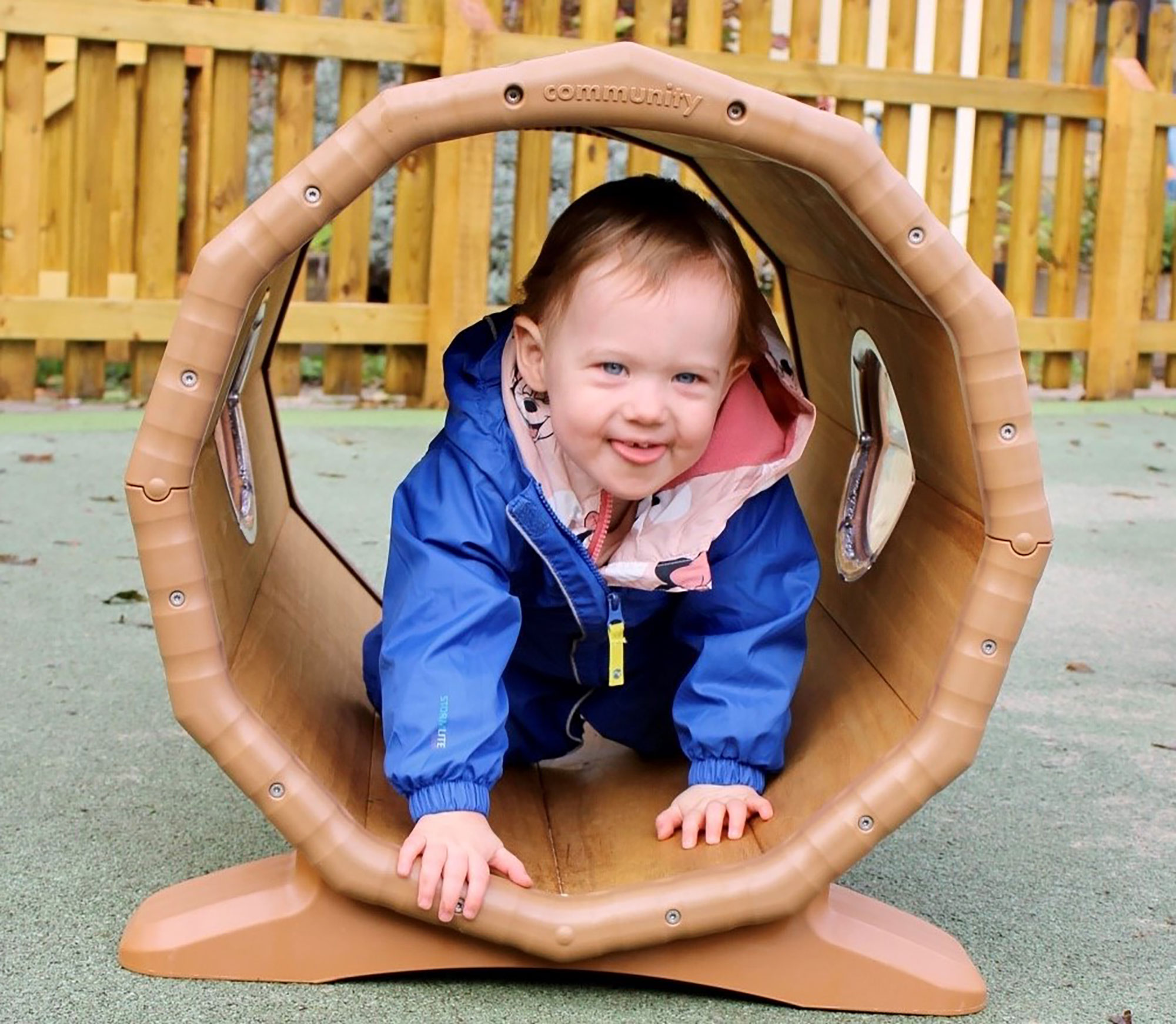 Acorns Nurseries A toddler in a blue jacket crawls through a play tunnel outdoors at Duffryn, with a wooden fence and scattered acorns in the background.