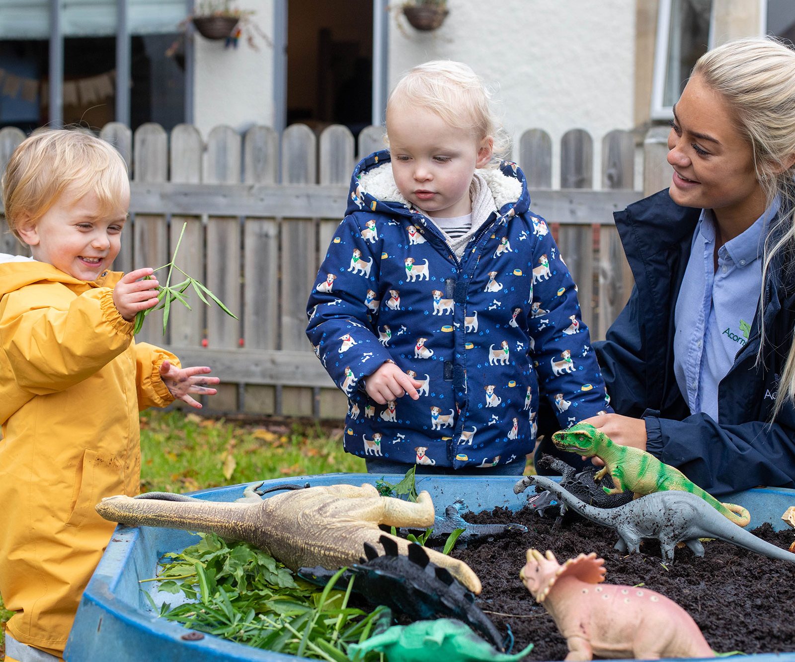 Acorns Nurseries Two toddlers and a woman interact with toy dinosaurs in an outdoor play area, surrounded by plants, acorns scattered about, and a wooden fence in Whitchurch.