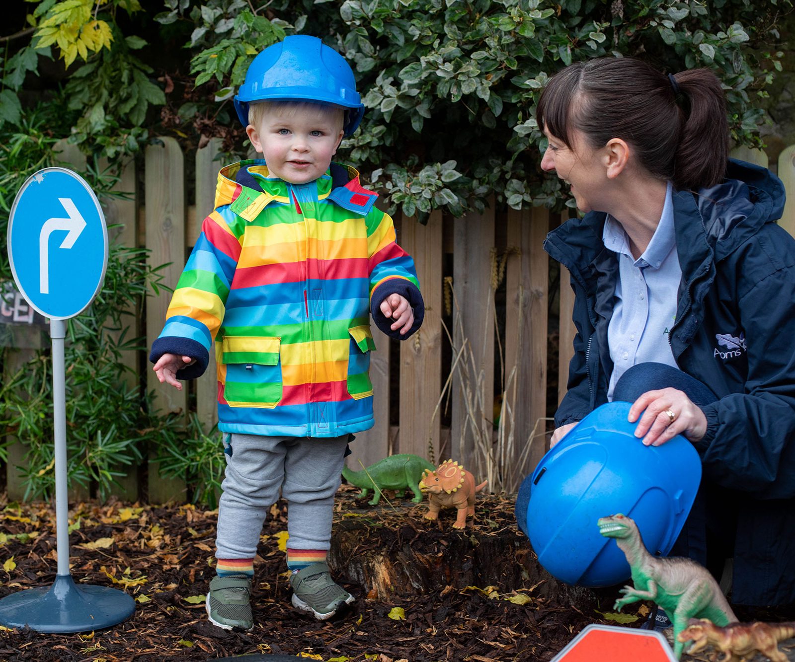 Acorns Nurseries A child in a rainbow coat and blue helmet stands by a right-turn sign in Whitchurch. An adult smiles, holding an Acorns helmet beside the child.