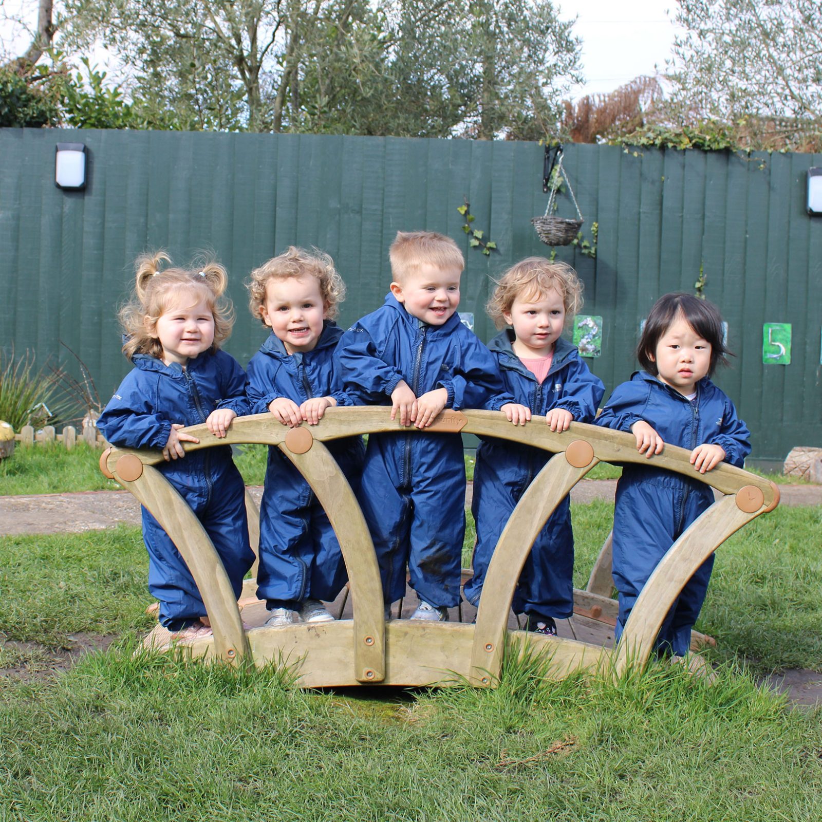 Acorns Nurseries Five children in blue outfits stand on a small wooden bridge in a grassy play area, watched over by the green fence. Nearby, acorns scatter the ground like nature's tiny treasures.