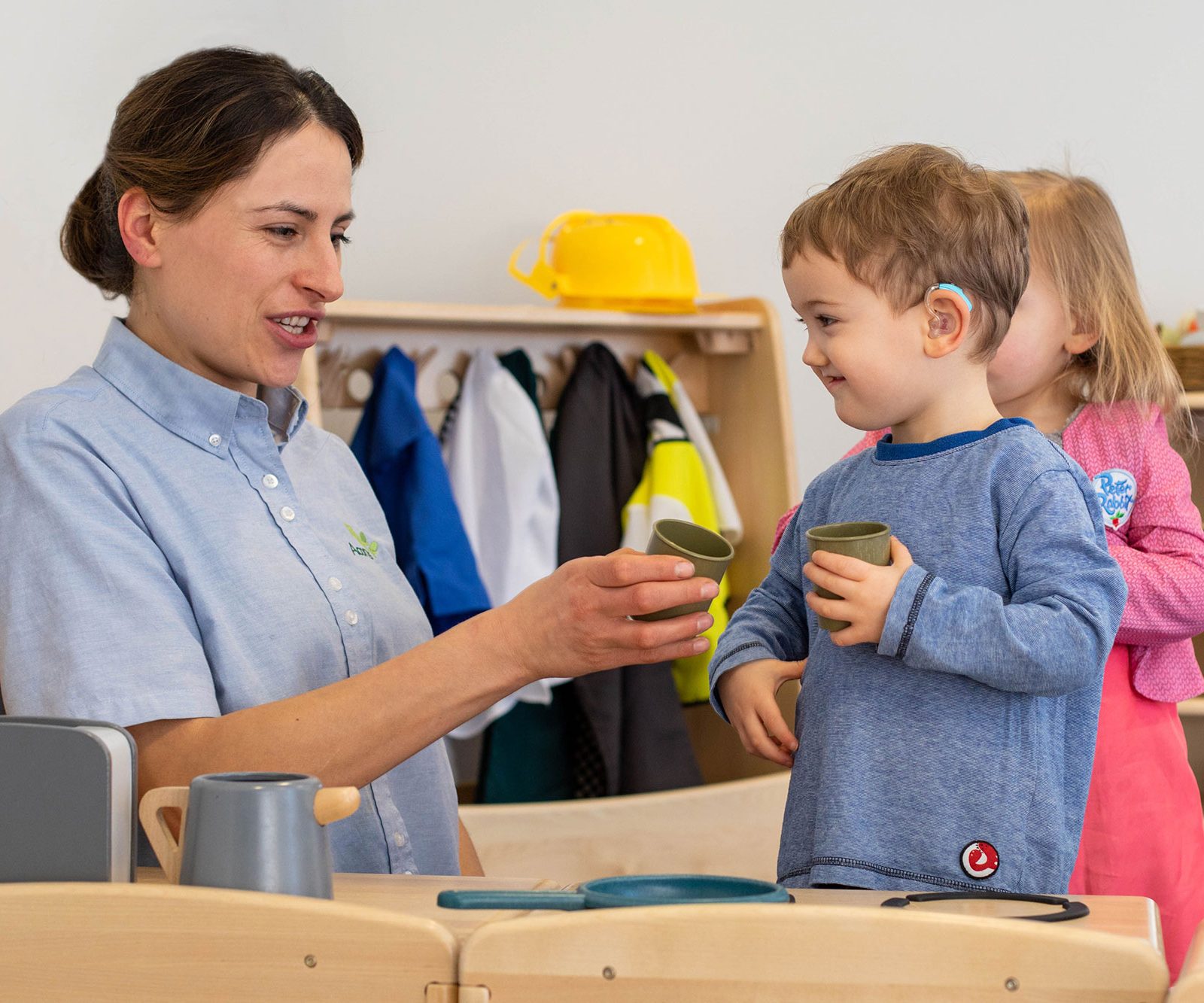 Acorns Nurseries In a vibrant classroom setting, reminiscent of a workplace nursery, a woman and two children joyfully play with toy cups. The boy eagerly joins in the fun, his hearing aid ensuring he's part of every laugh and whisper.