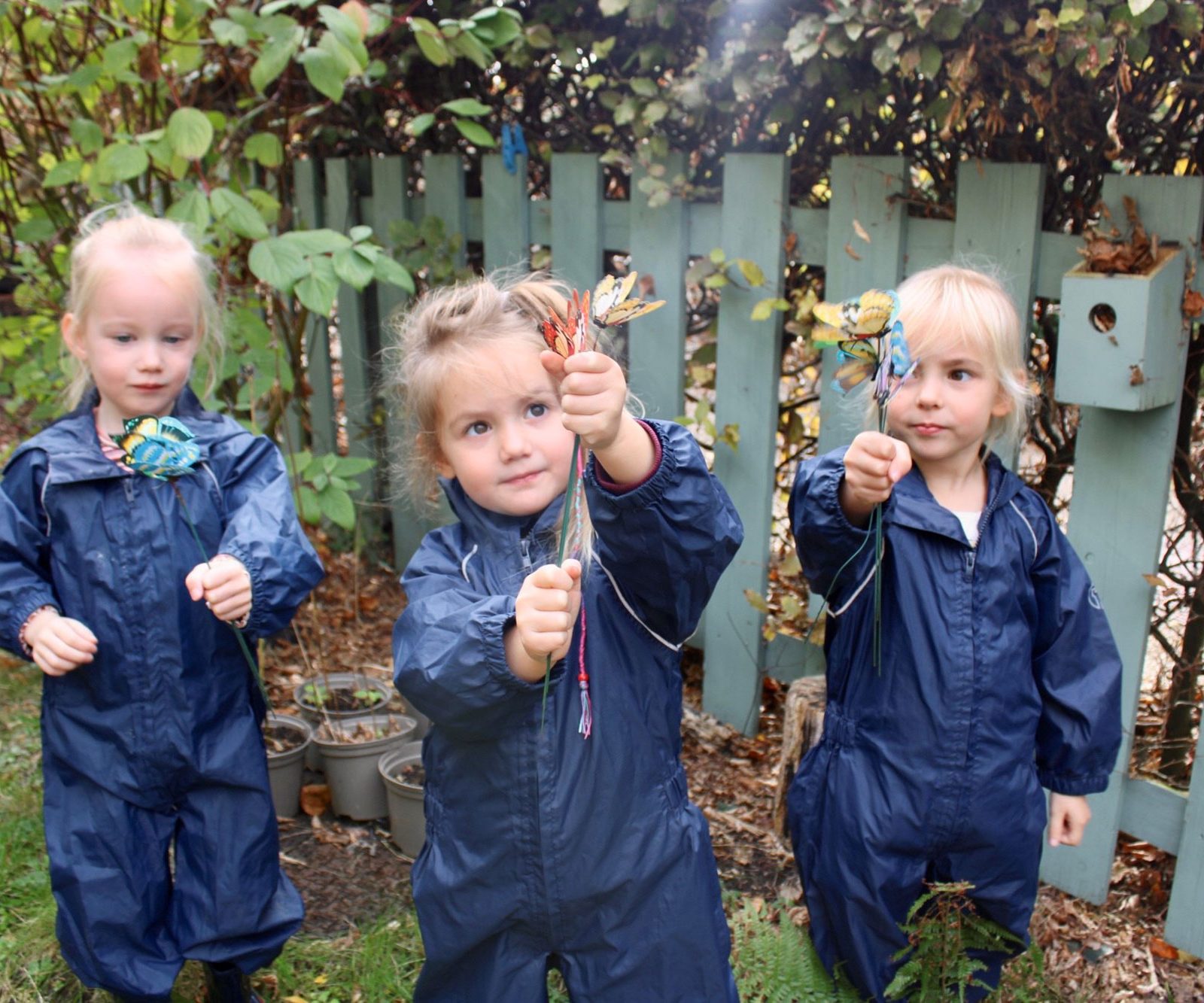 Acorns Nurseries Three children in blue overalls, members of the Acorns Aviva workplace nursery, proudly hold their crafted items as they stand in front of a wooden fence within the vibrant garden area.