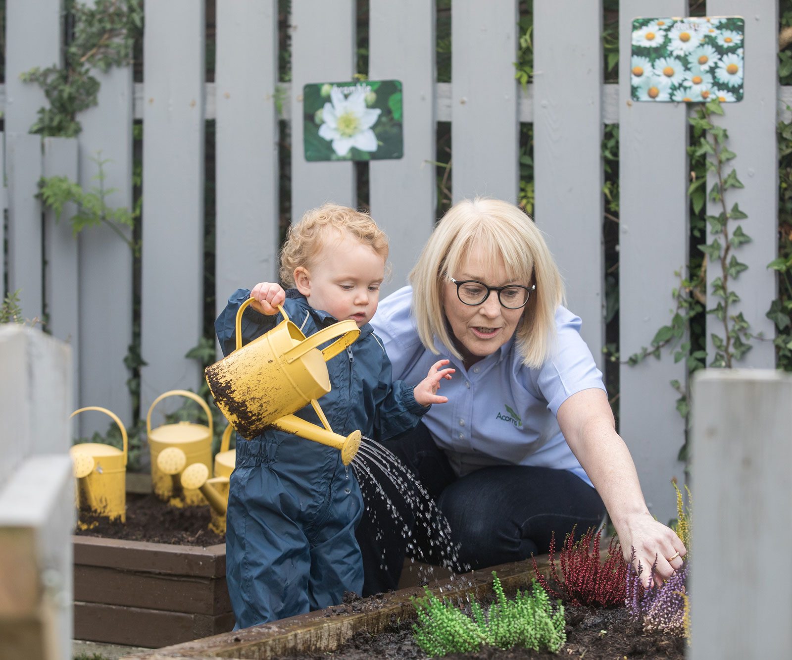 Acorns Nurseries A baby in a blue rain suit waters plants under adult guidance in a garden, with yellow watering cans scattered around.