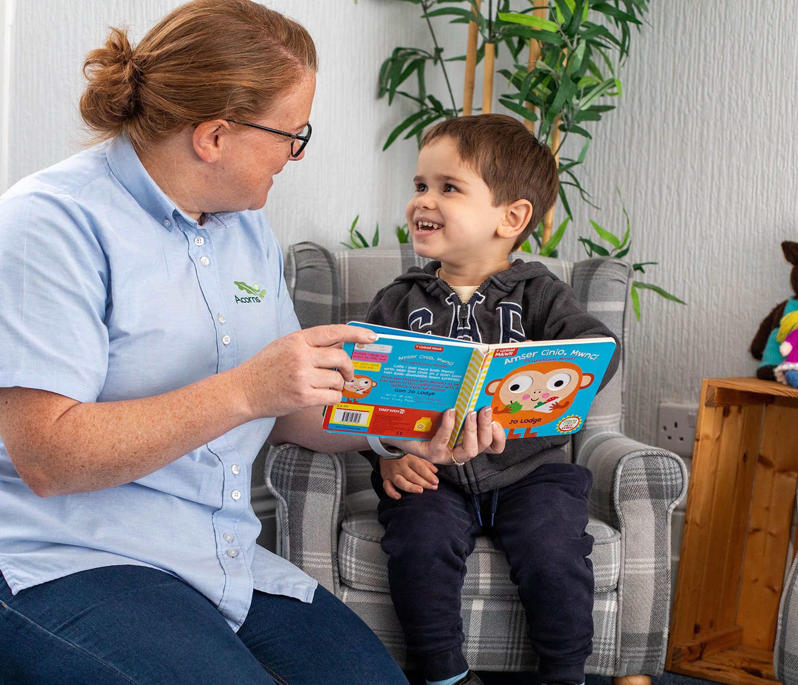 Acorns Nurseries An adult and a child sit on chairs in the cozy nook of Acorns House, smiling at each other while the adult holds an open children's book.