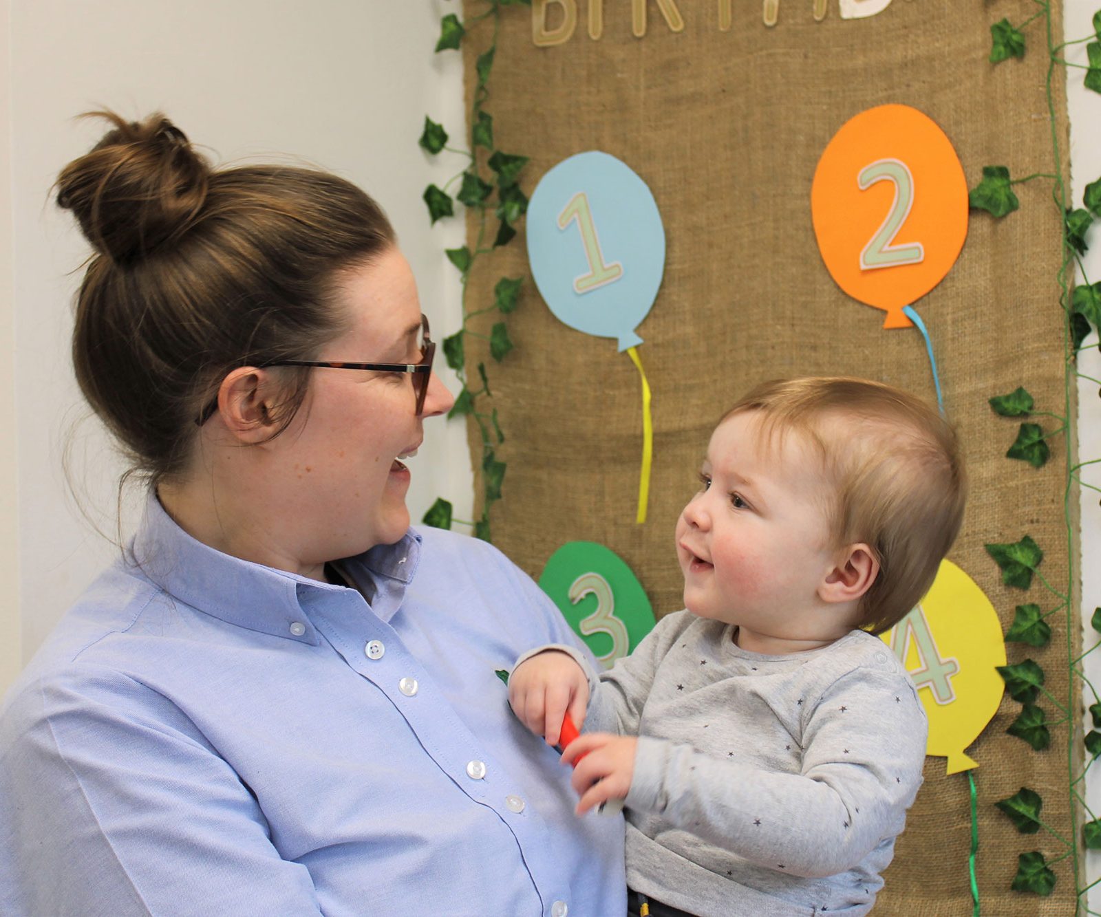 Acorns Nurseries A woman smiles and holds a toddler in front of a decorative wall with colored balloons numbered 1 to 4, capturing the joy of the Workplace Nursery celebration.