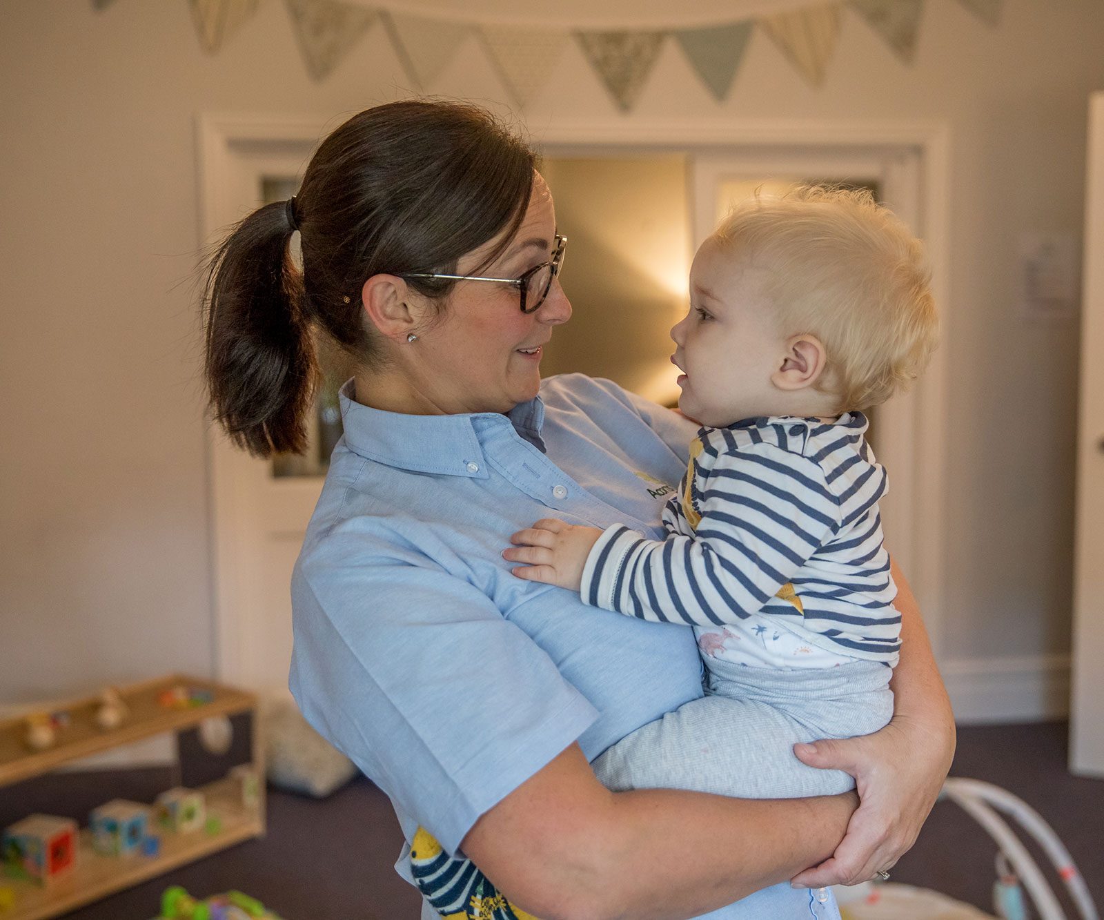 Acorns Nurseries A woman in a blue shirt gently cradles a baby in a striped shirt amidst playful acorns, toys, and a colorful banner adorning the room.