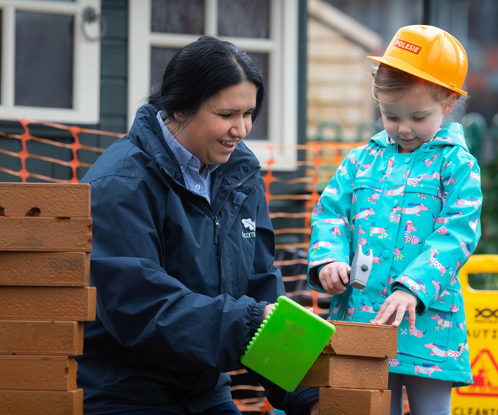 Acorns Nurseries An adult and child at the Workplace Nursery are building with toy bricks; the child wears a construction helmet and a colorful coat, embodying the spirit of imagination fostered by Acorns ONS.