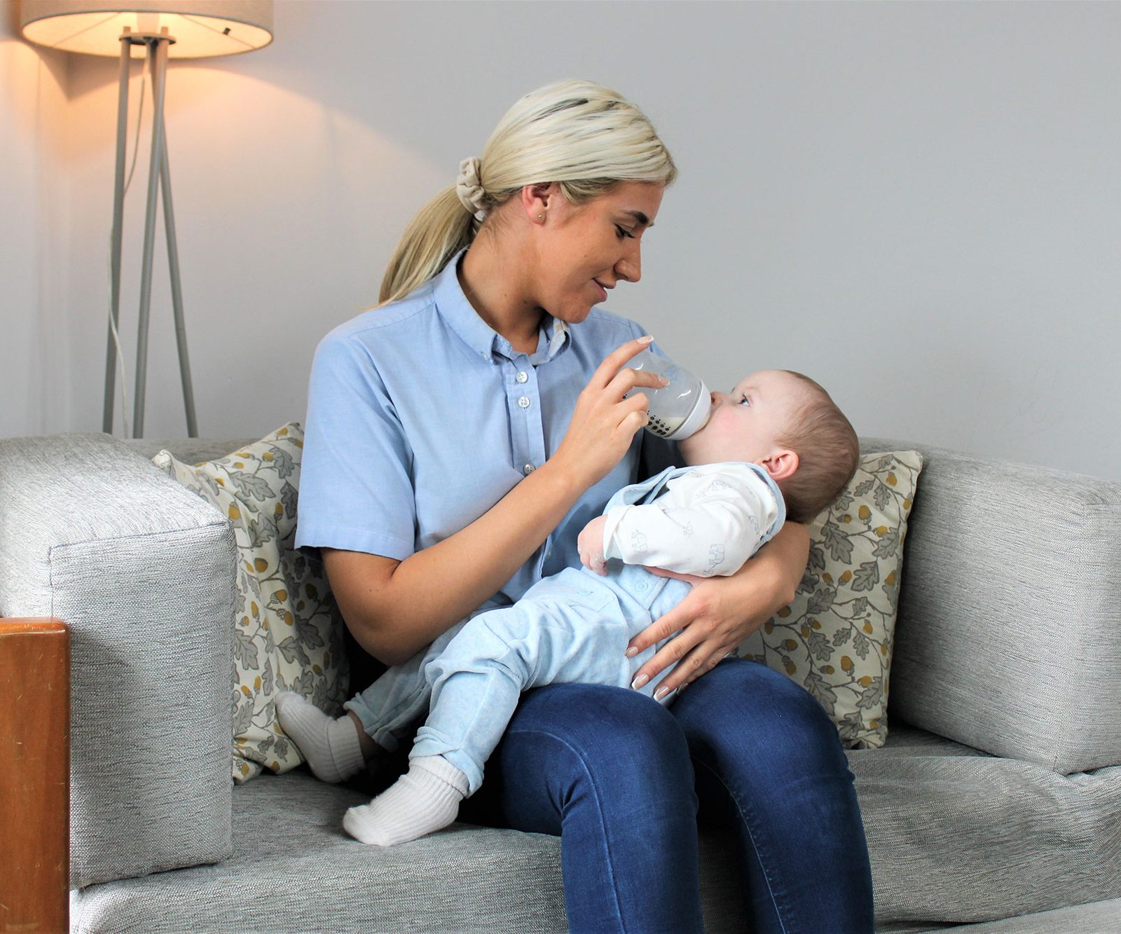 Acorns Nurseries A person with blonde hair sits on a sofa in Parc Nantgarw, bottle-feeding an infant dressed in light blue. A lamp glows softly in the background, casting a warm light reminiscent of acorns scattered on an autumn day.