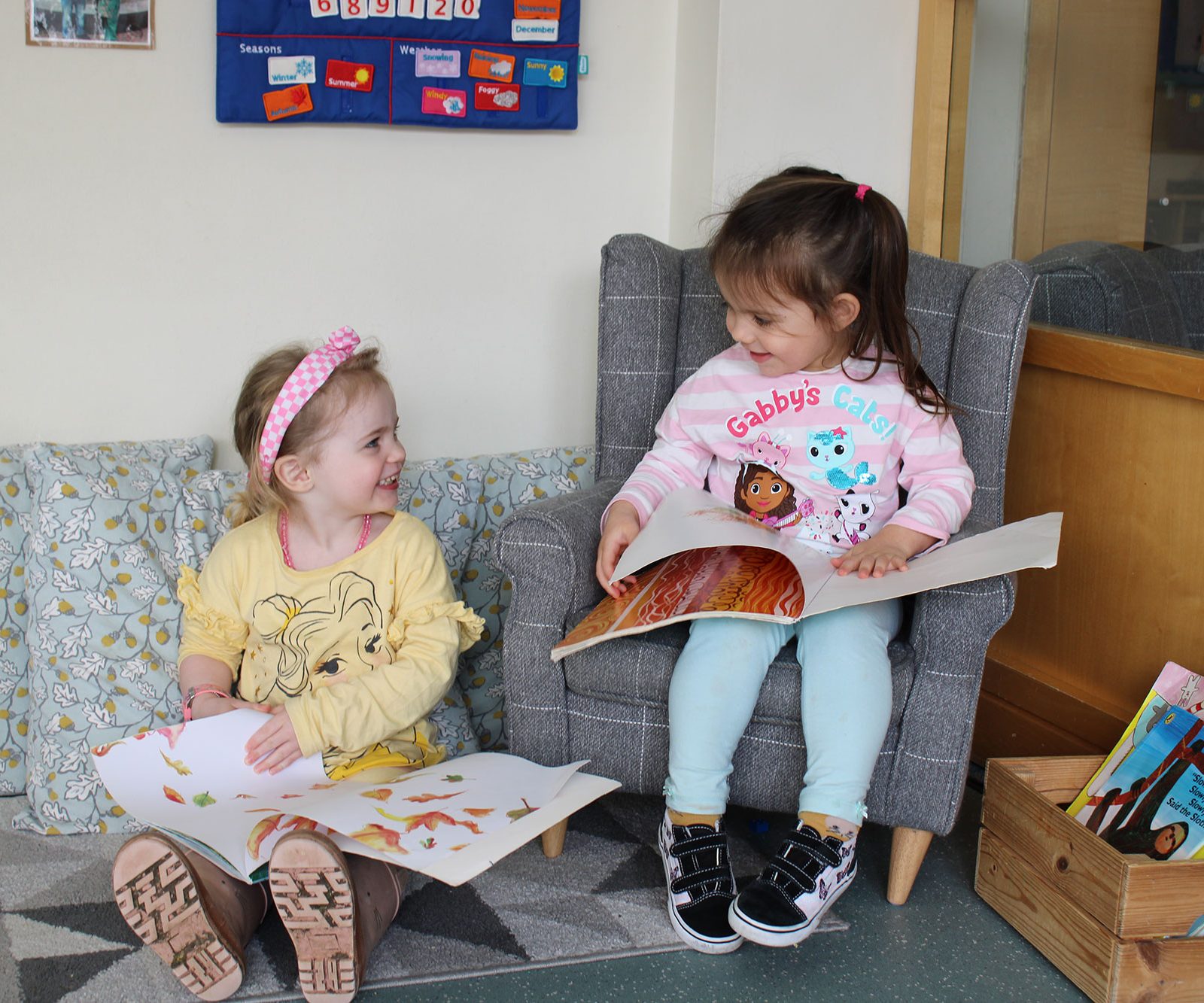 Acorns Nurseries Two young children sit on a couch, each holding a book, in a cozy corner reminiscent of an Aviva workplace nursery. A small bookshelf brimming with stories stands nearby, nurturing tiny acorns of imagination and growth.