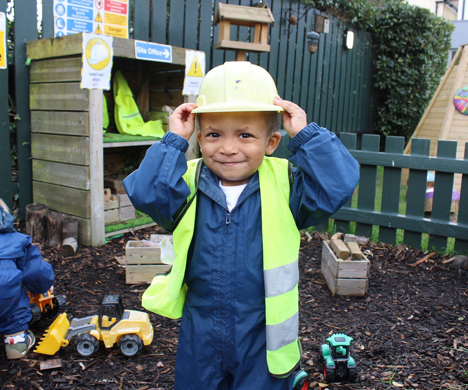 Acorns Nurseries A child wearing a yellow hard hat and safety vest beams with joy while playing in the Baby Acorns construction-themed playground.