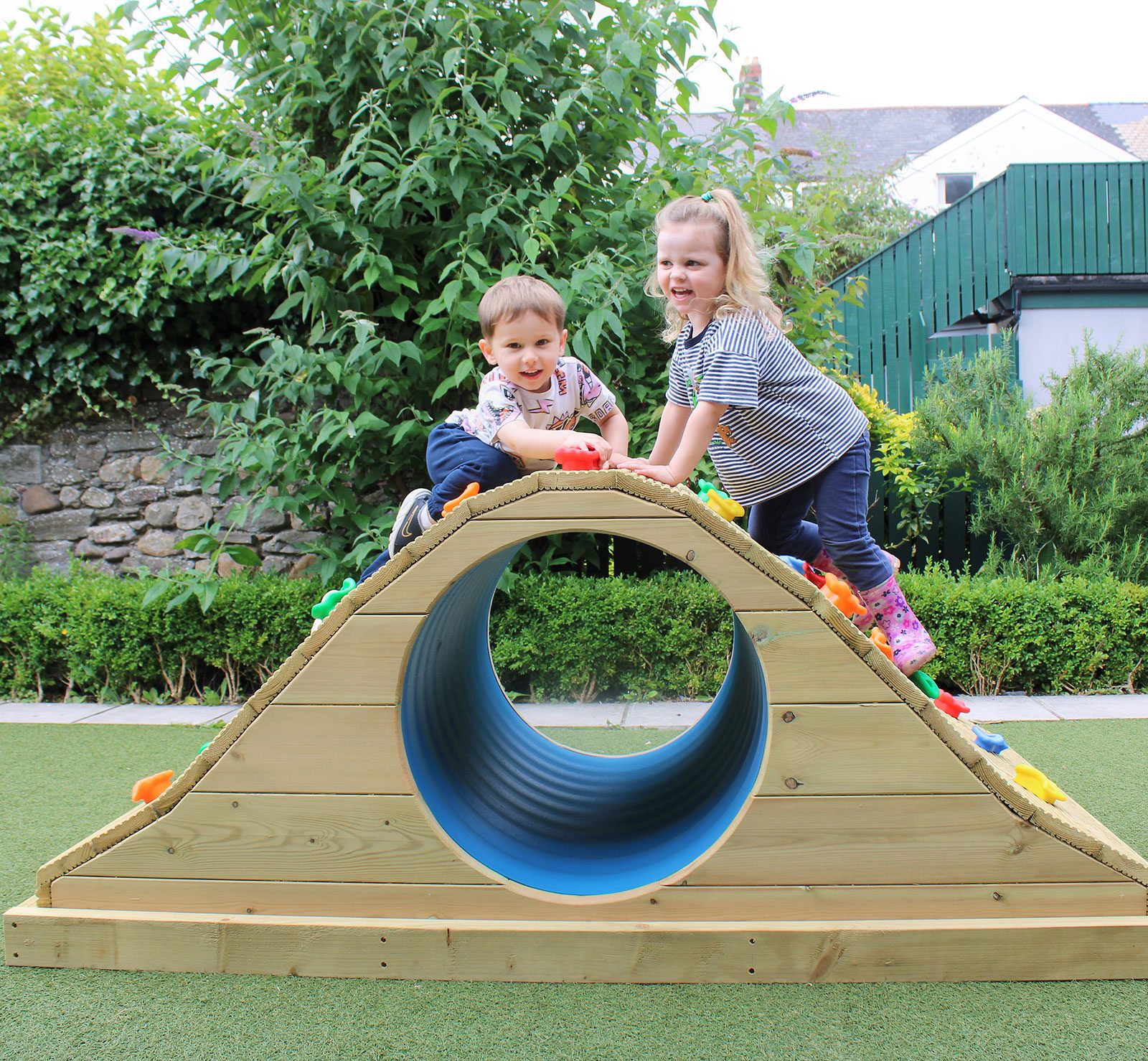 Acorns Nurseries Two children climb on a wooden play structure shaped like a bridge with a tunnel passage, as the sun beams down on Acorns House's vibrant outdoor playground.