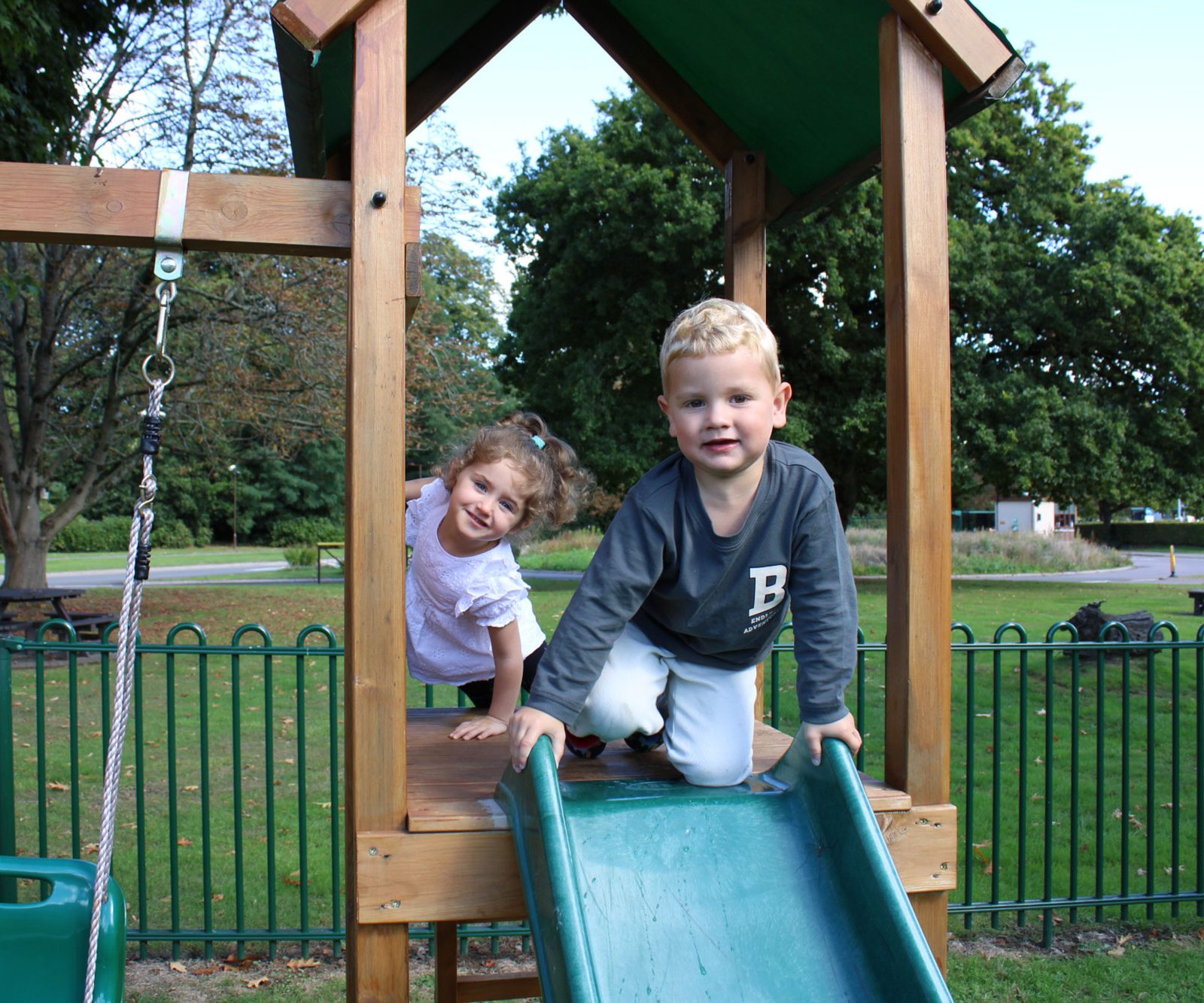 Acorns Nurseries Two children giggle as they slide down a wooden slide in a park, nestled among trees and bordered by a metal fence. The scene is reminiscent of the warmth found in an Acorns ONS workplace nursery, where exploration and play go hand-in-hand with curiosity and joy.