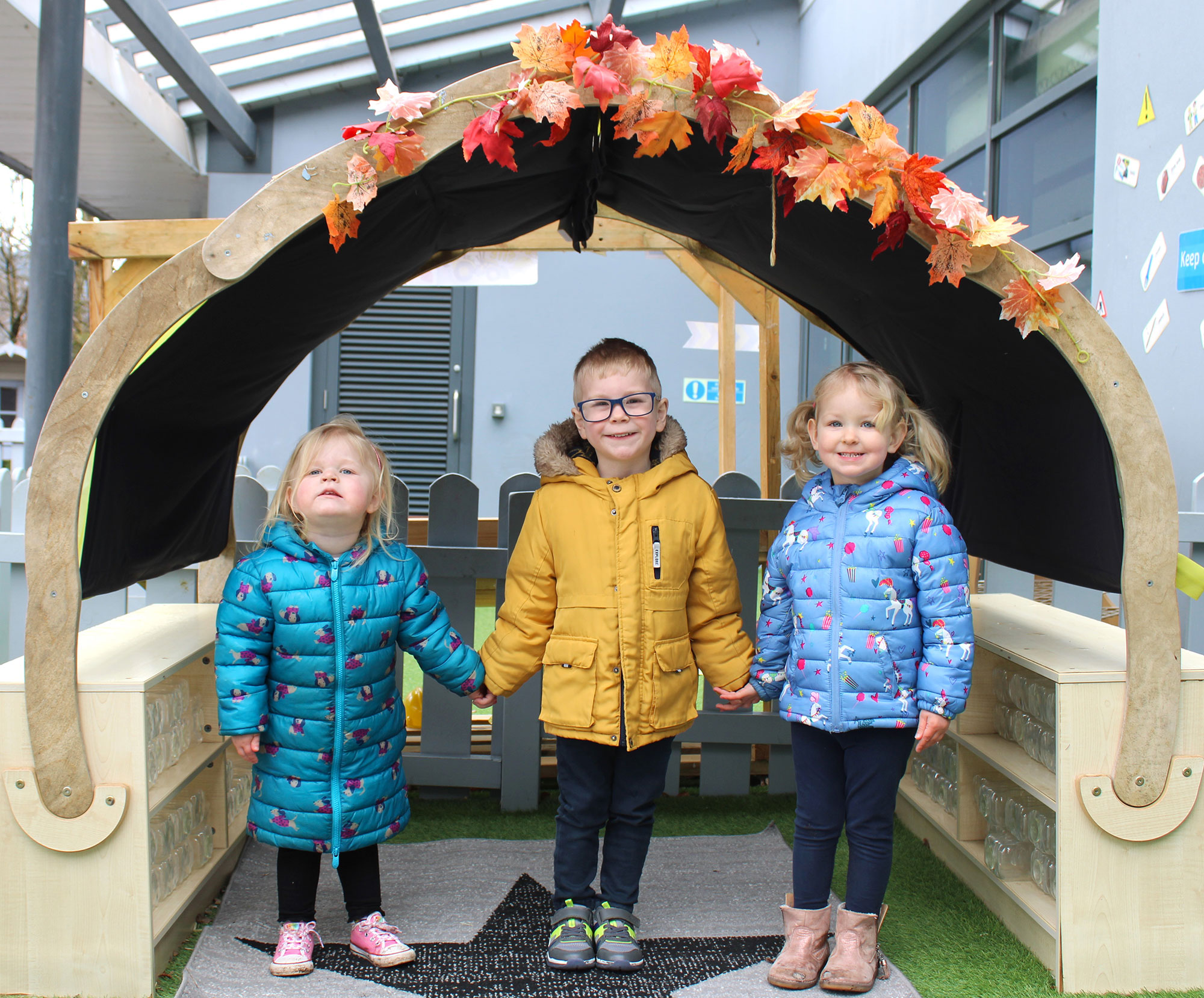 Acorns Nurseries Three children in winter coats hold hands under a decorative arch adorned with autumn leaves and scattered acorns, reminiscent of a charming scene in Parc Nantgarw.